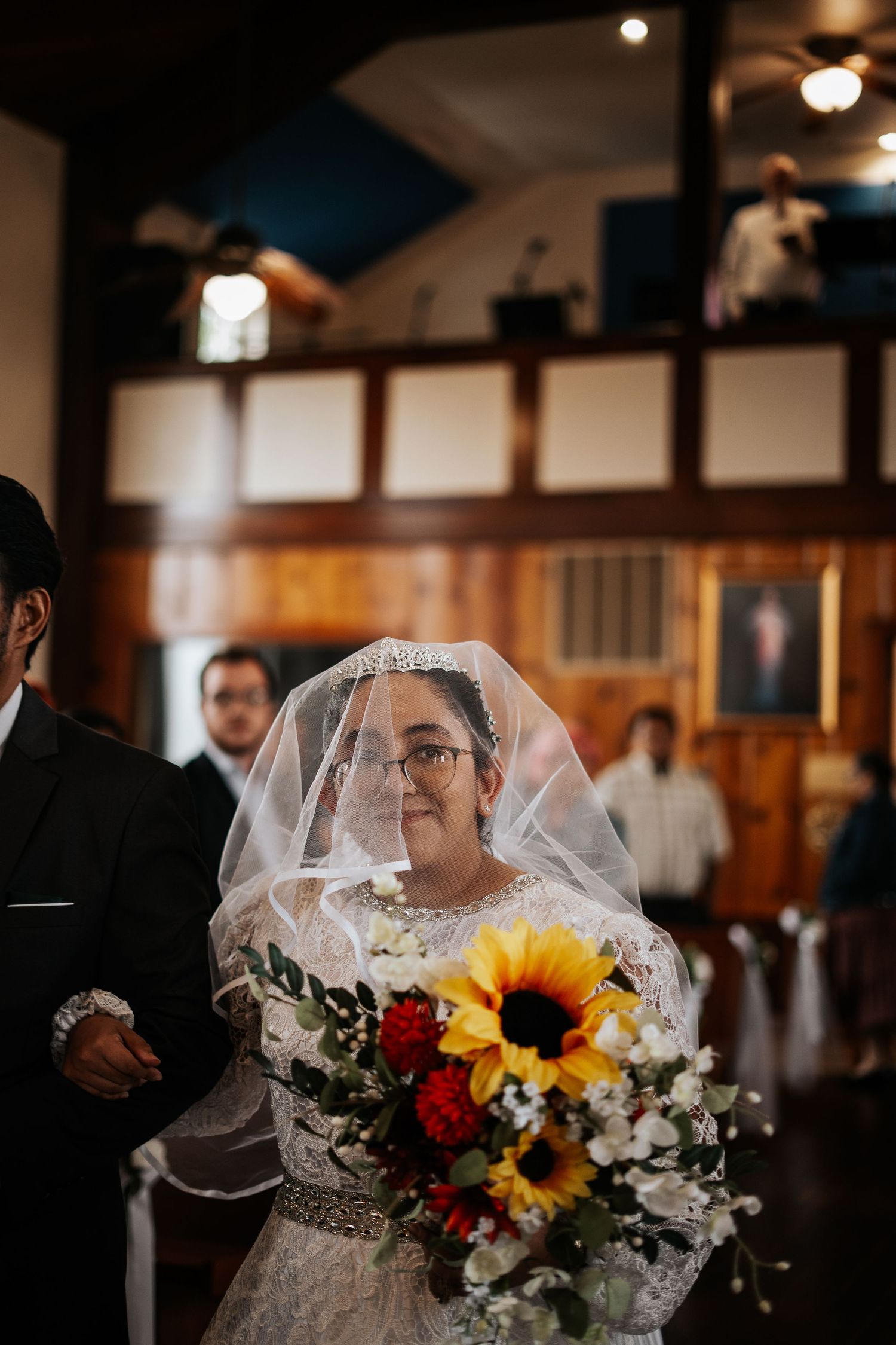 Bride walking down church aisle in lace gown and veil, holding sunflower and red rose bouquet at wedding ceremony.