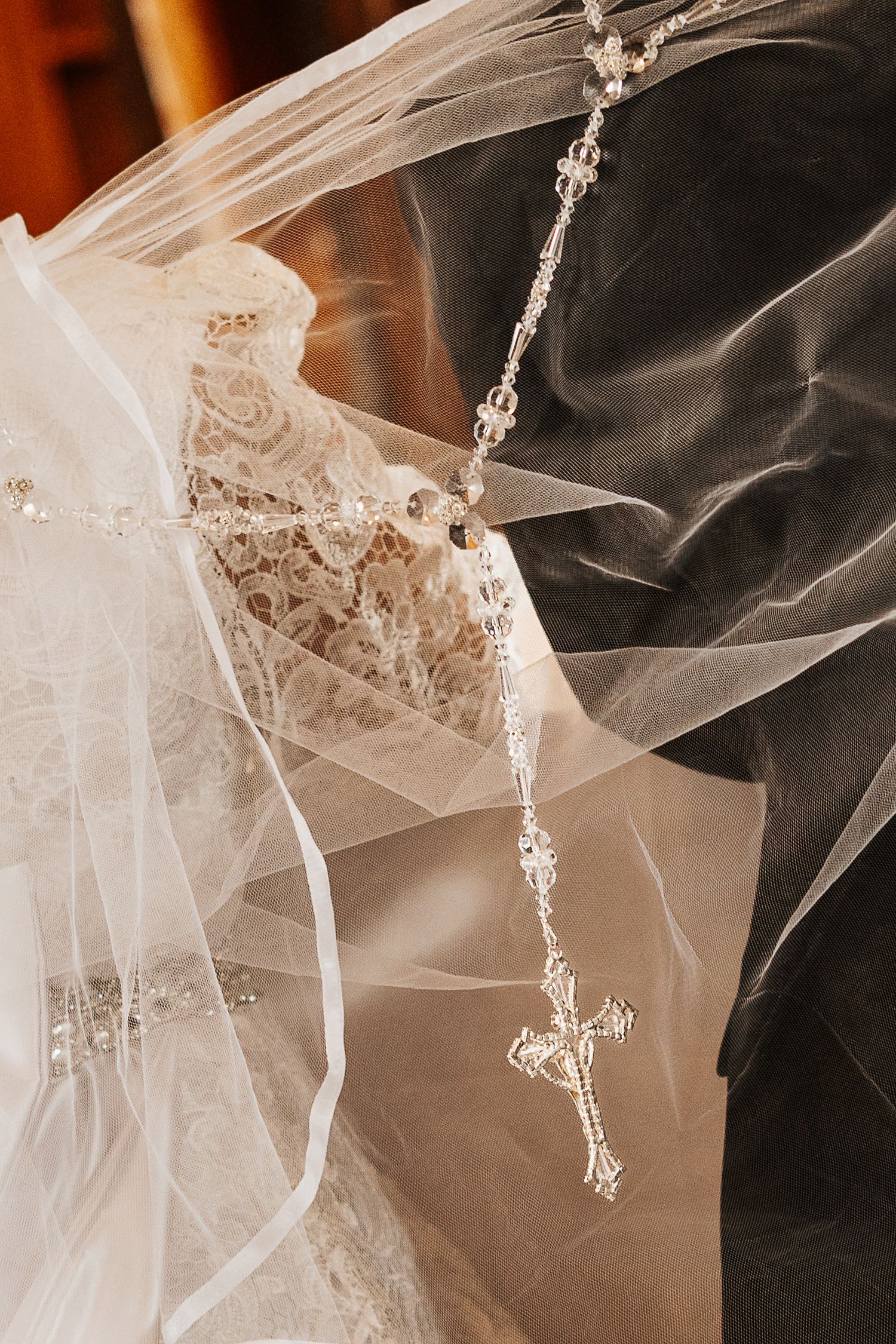 Close-up of bride and groom embracing, showcasing lace wedding dress, tulle veil, and crystal rosary cross detail.