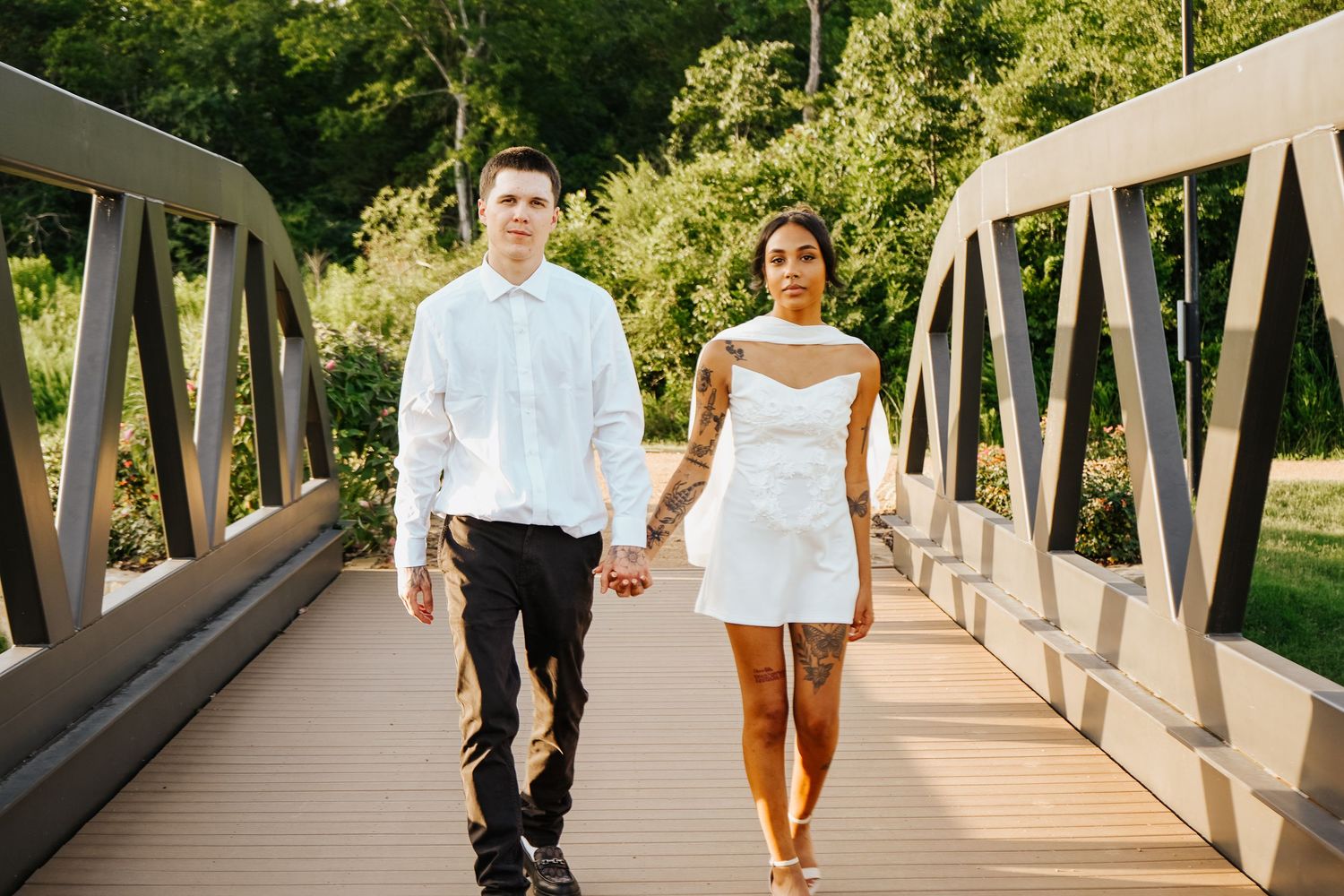 Engaged couple walking hand in hand across modern bridge surrounded by lush greenery, woman with tattoos.