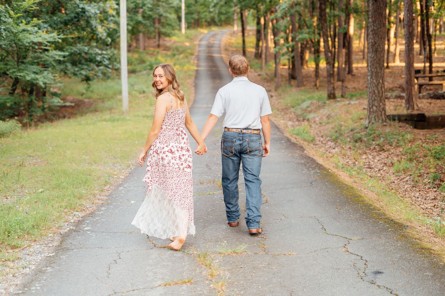 Couple walking hand in hand down a tree-lined path, woman in floral maxi dress and man in jeans.
