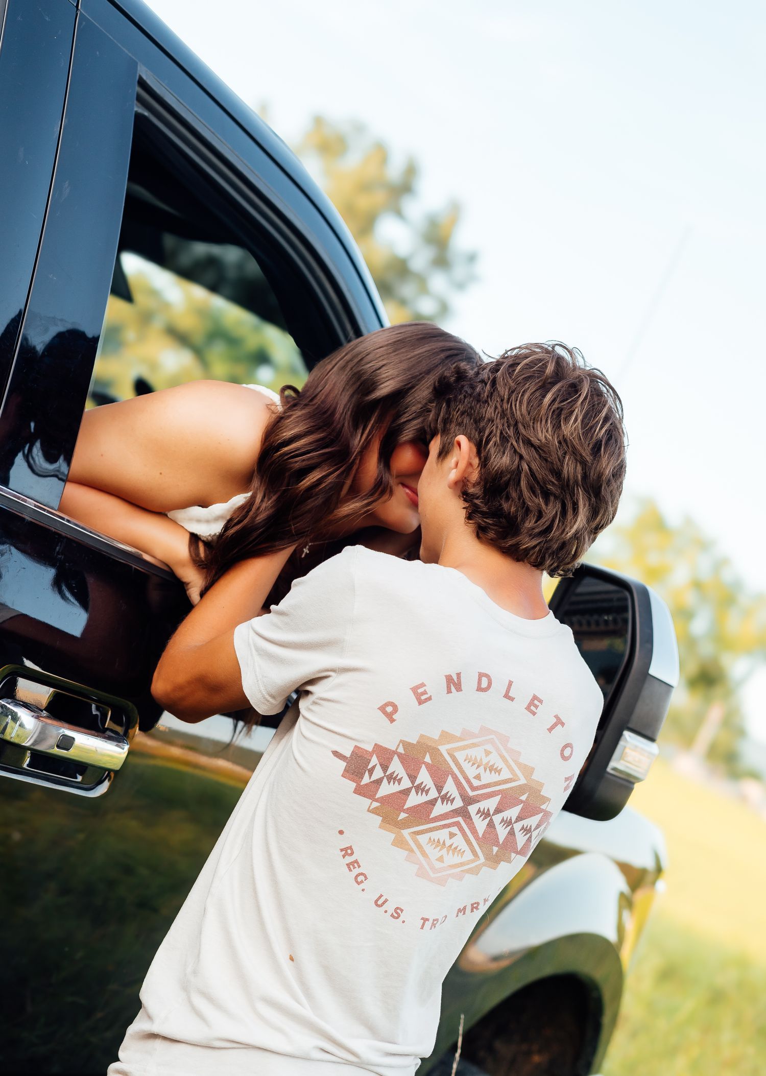 Young couple sharing a kiss near a dark SUV outdoors with green trees in the background.