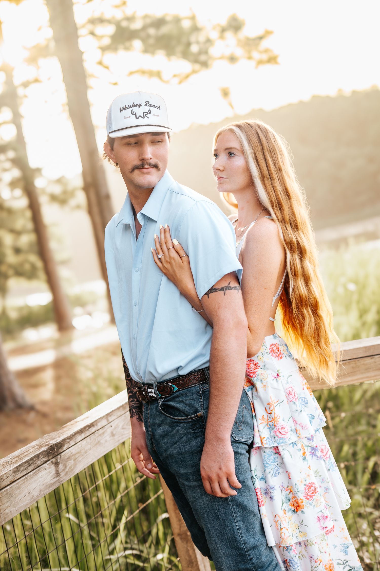 Couple posing by wooden fence at golden hour, man in white cap and blue shirt with tattooed woman in floral dress.