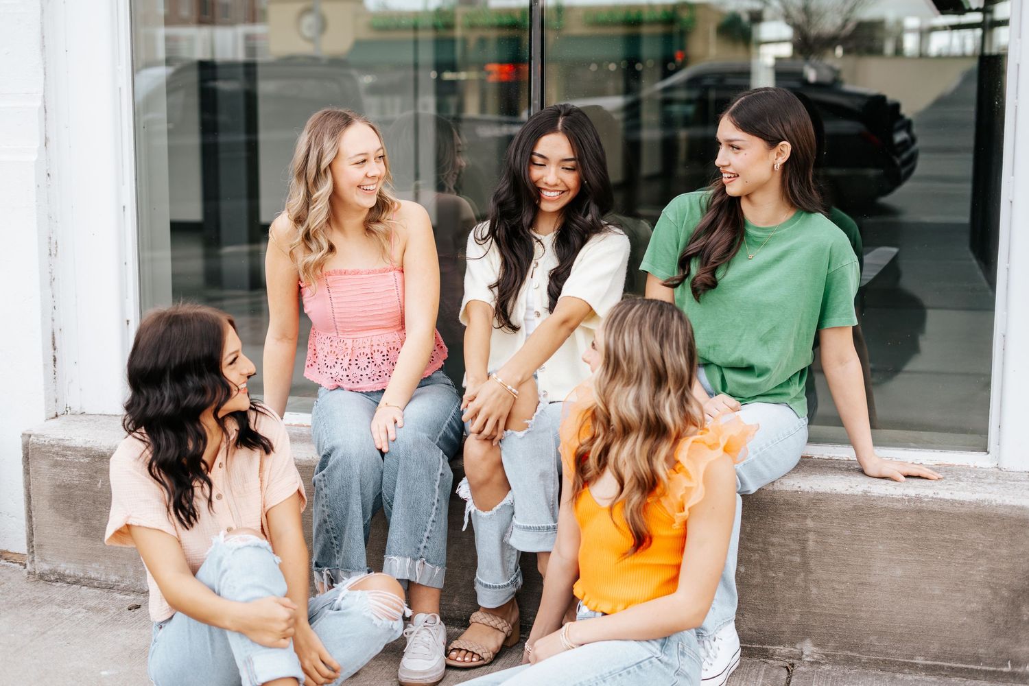 Group of five young women in casual colorful tops and jeans sitting and chatting outside a storefront.