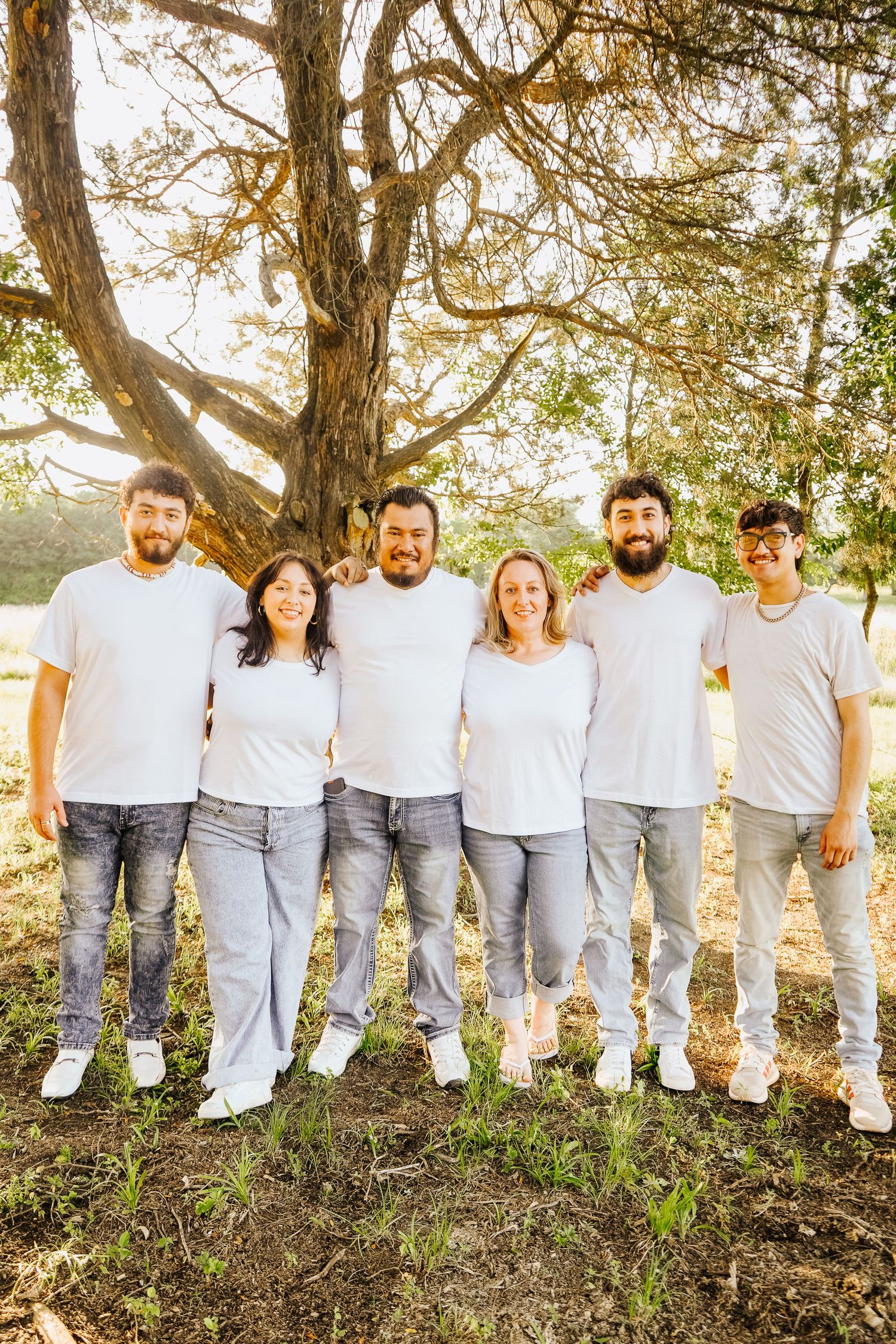 Family of five posing in matching white tees and jeans under a large tree outdoors.