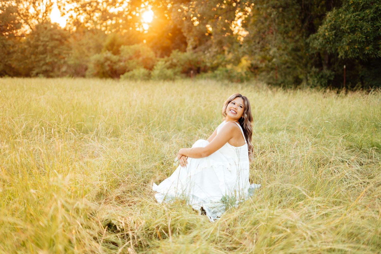 Happy young woman in white dress sits smiling in tall grass field during golden sunset.