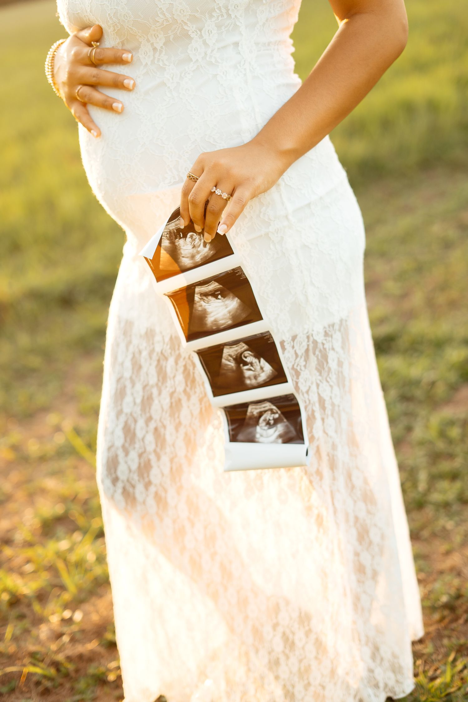 Pregnant woman in white lace dress holding ultrasound photos outdoors at golden hour.