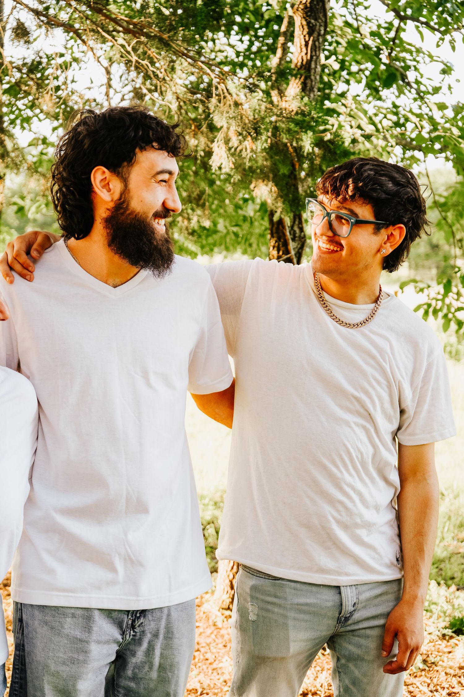 Two bearded brothers in white tees smiling and embracing outdoors under green trees.