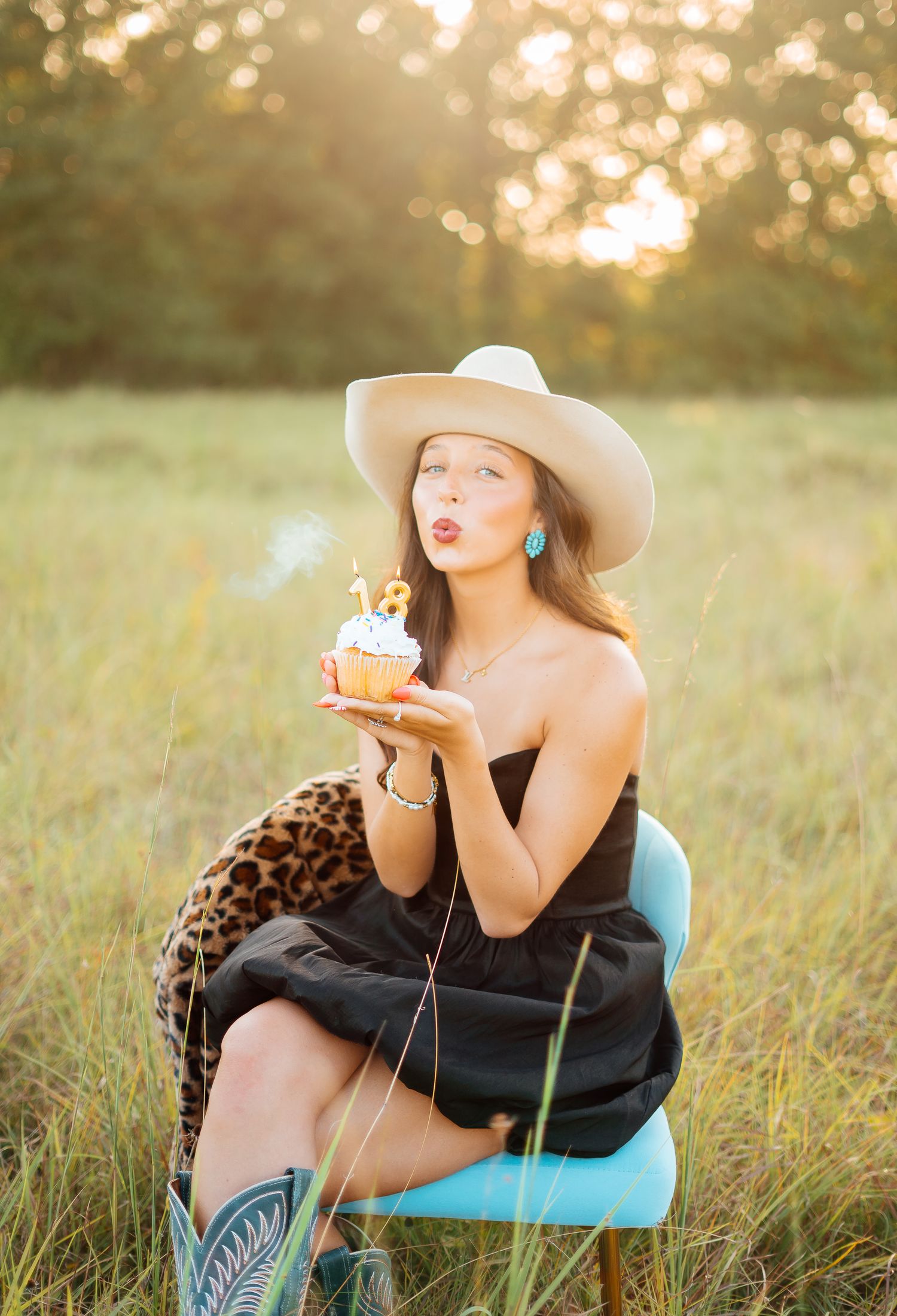 Young woman in black dress blows out candle on cupcake while sitting in sunny meadow.
