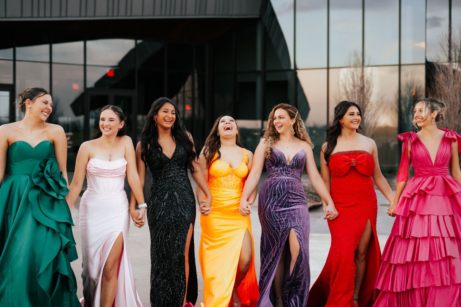 Seven women in colorful formal prom dresses laughing and walking together outside modern building.
