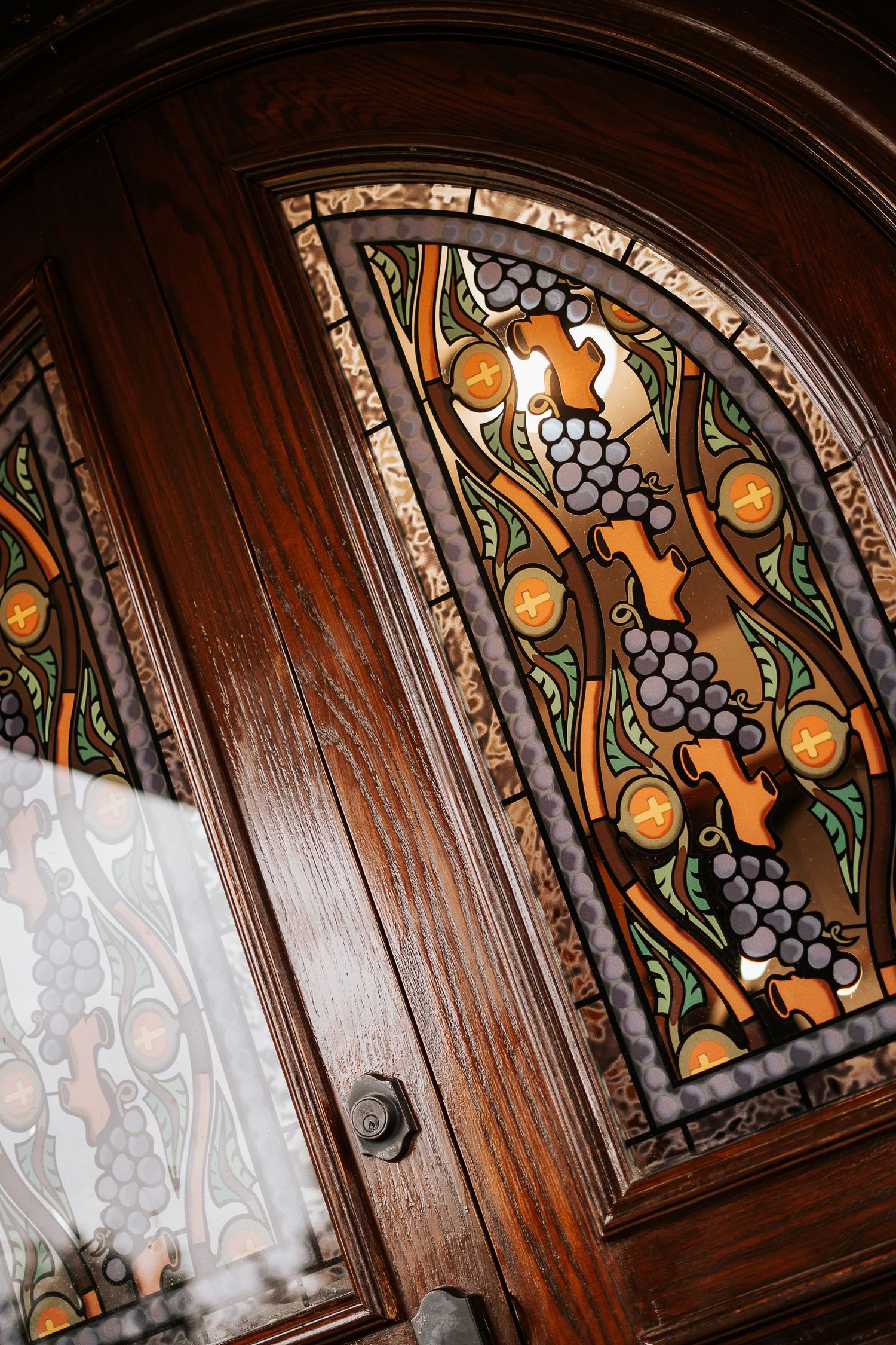 Ornate wooden door with arched stained glass panels featuring grape and floral motifs in rich colors.