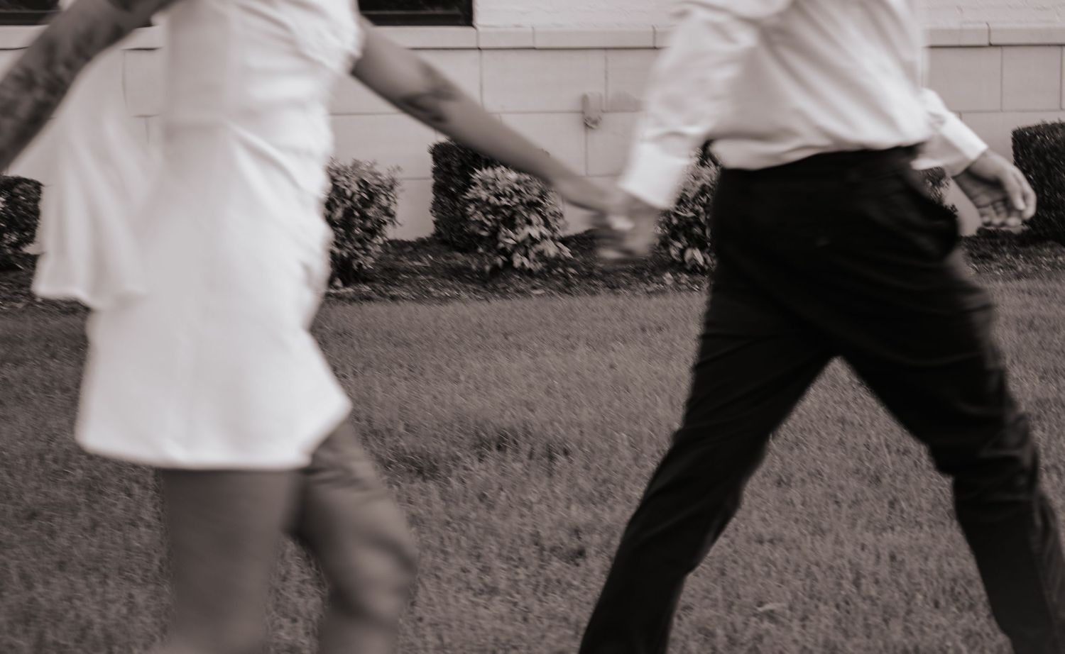 Black and white motion blur photo of bride in mini dress and groom holding hands walking across lawn.