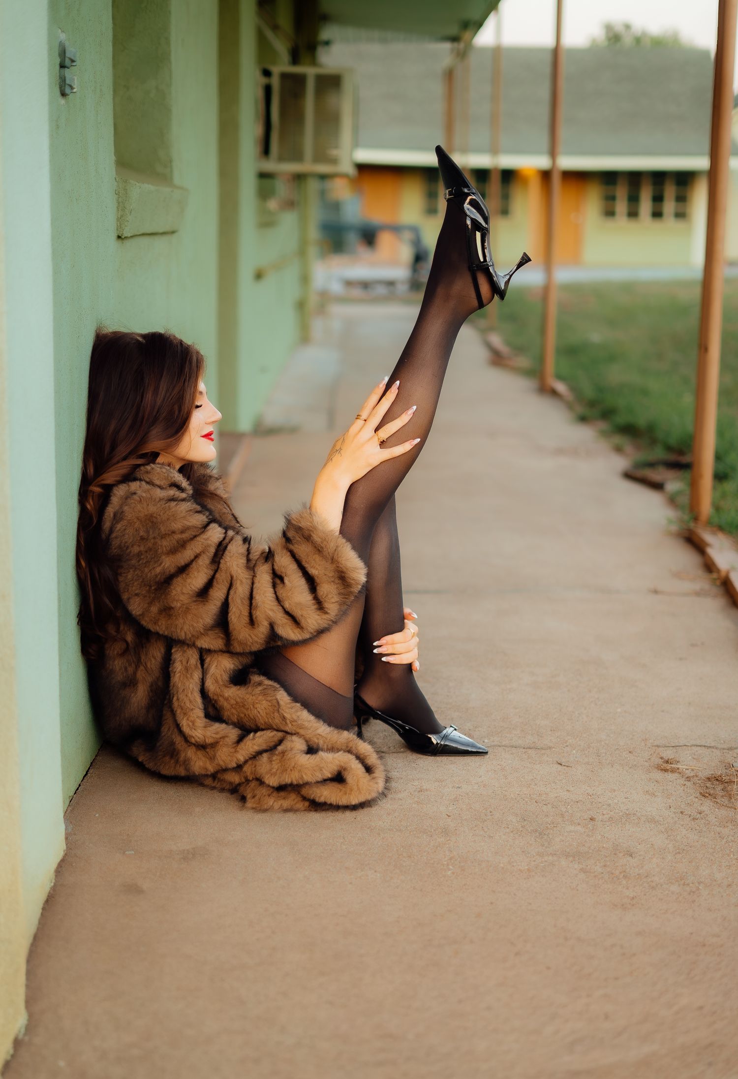 Woman in fur coat and black stockings posing playfully against a pastel motel wall.