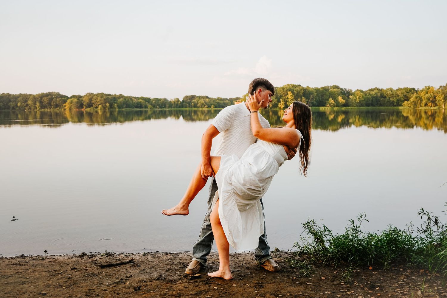 Man lifts woman in white dress for a romantic kiss near a serene lakeside at dusk.