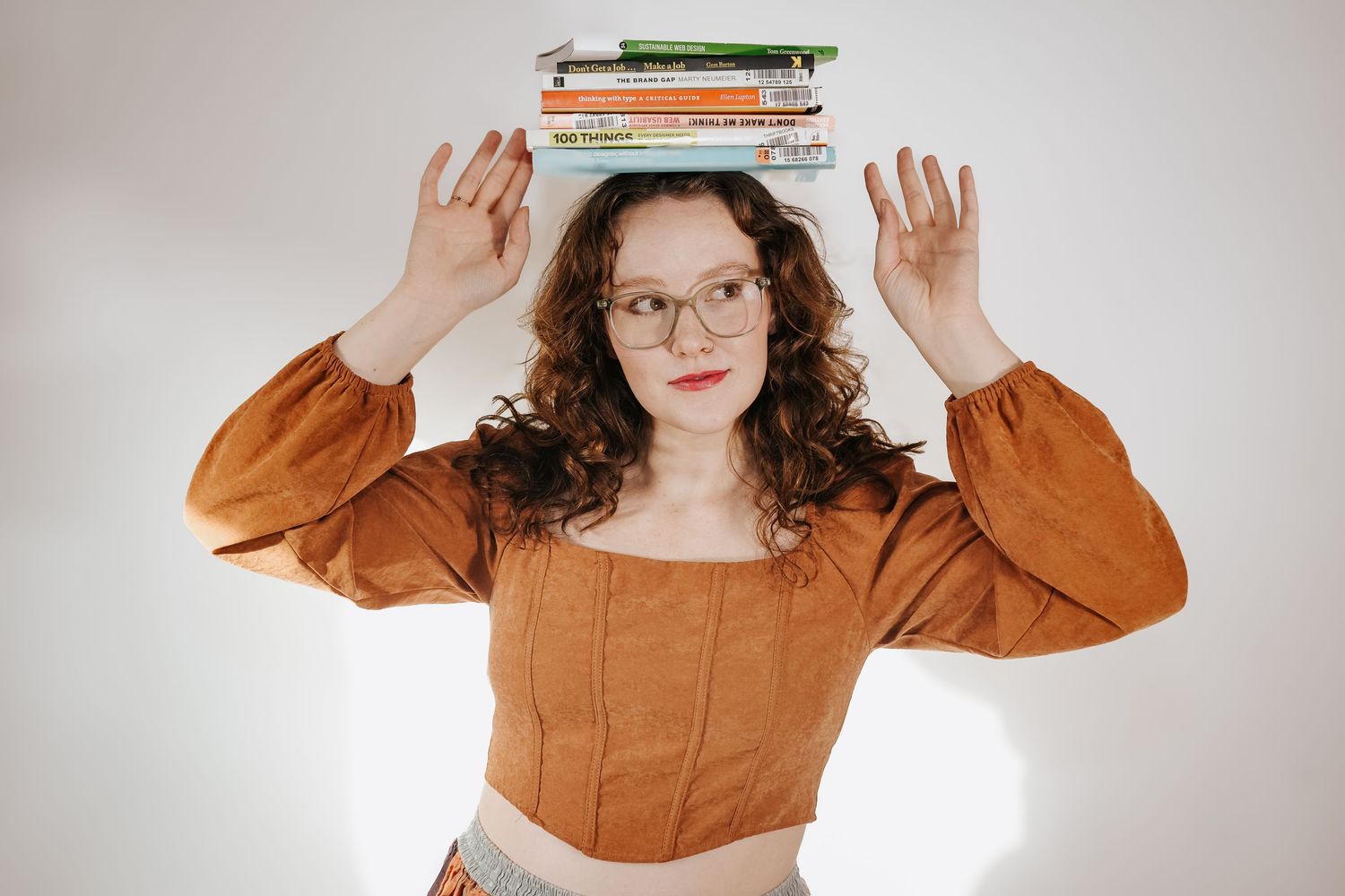 Young woman with curly hair and glasses balancing a stack of colorful books on her head against white background.