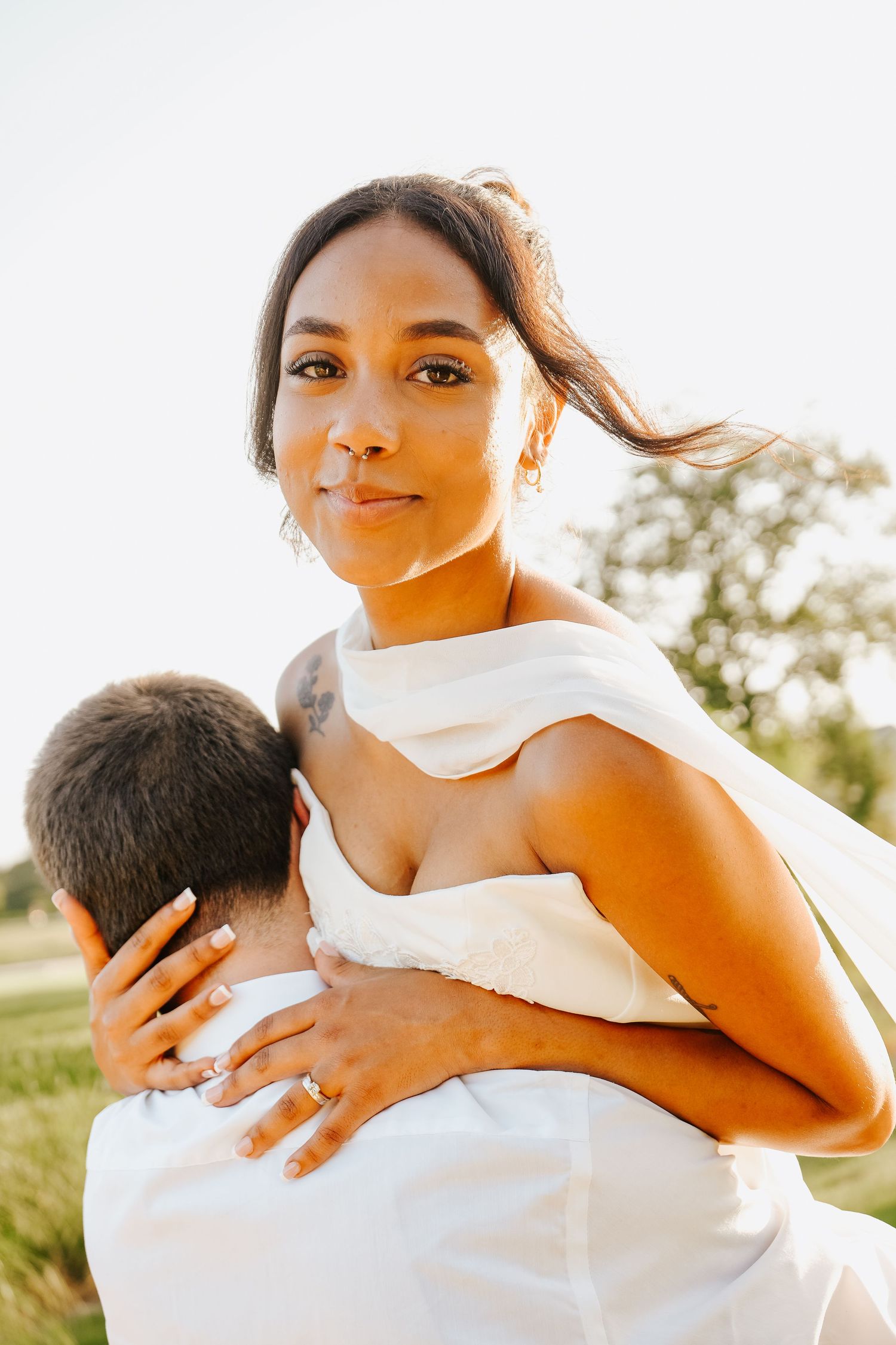 Bride in white halter dress with engagement ring embracing groom outdoors during golden hour portrait session.