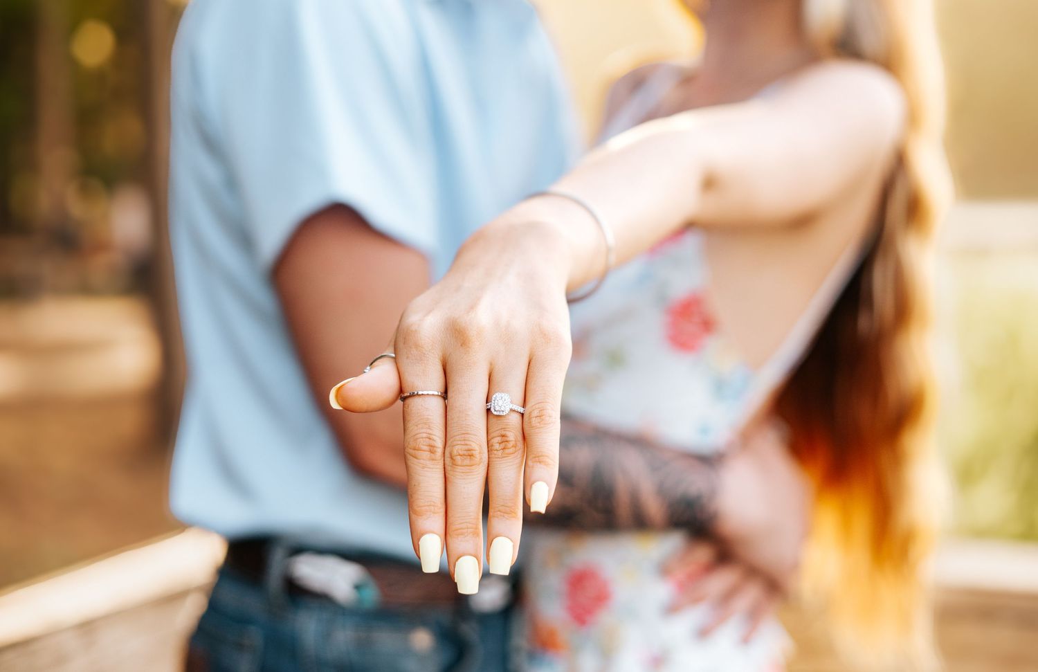 Close-up of woman showing diamond engagement ring while embracing partner in sunny outdoor setting.