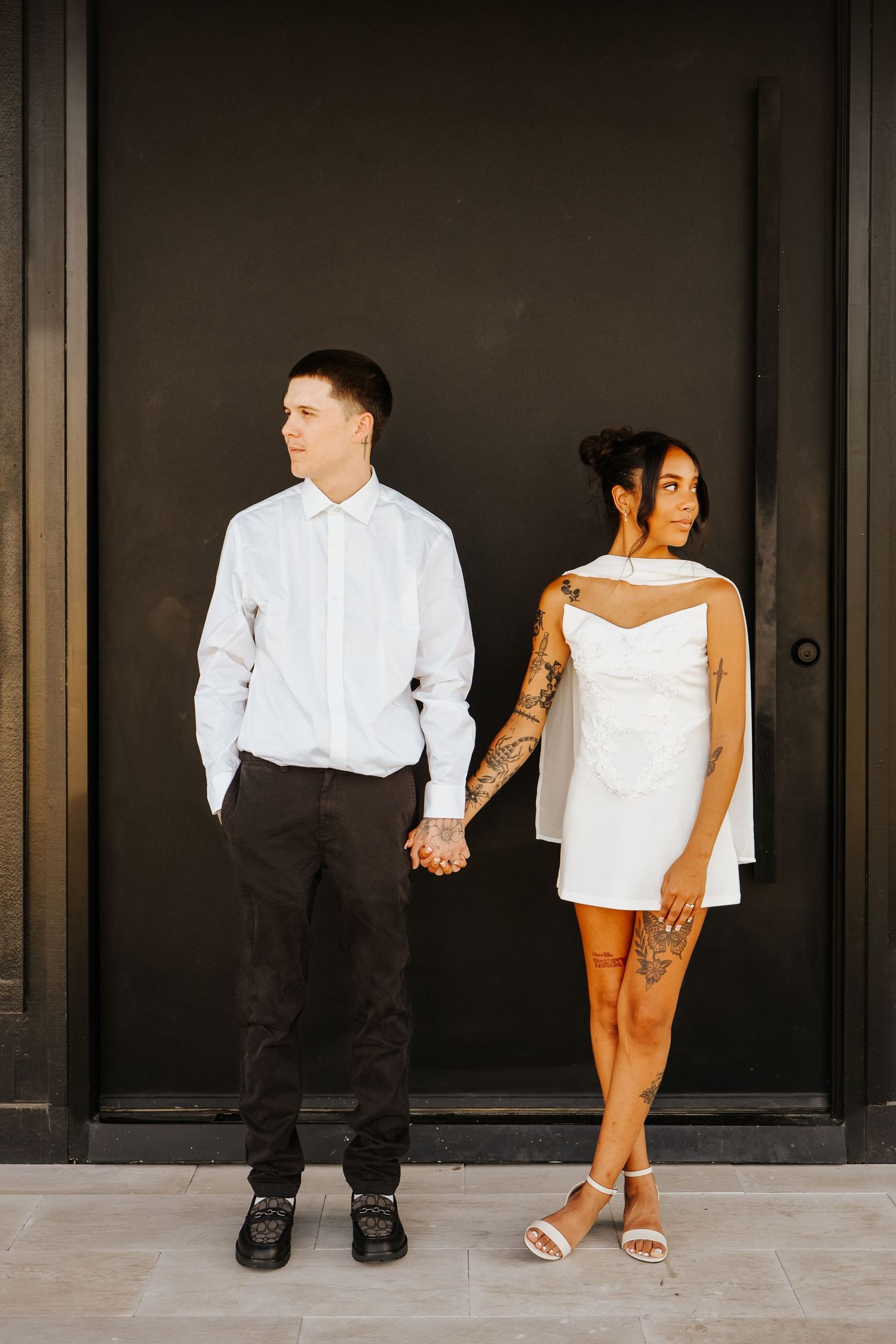 Engaged couple holding hands in front of black wall, woman in white dress with tattoos, man in white shirt.