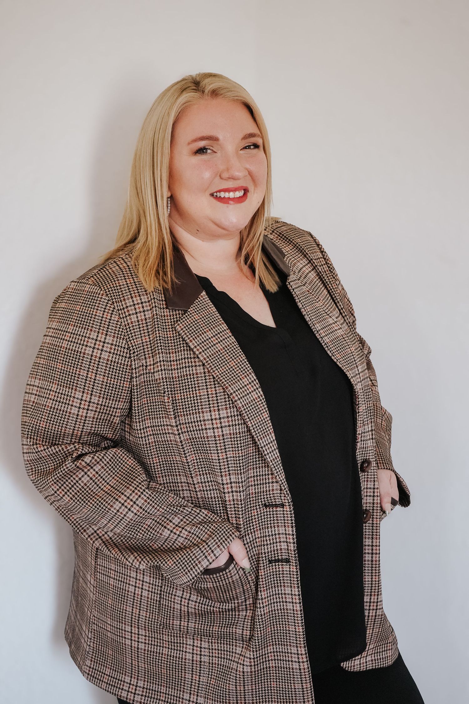 Smiling blonde woman in a plaid blazer over black outfit posing against a white wall.