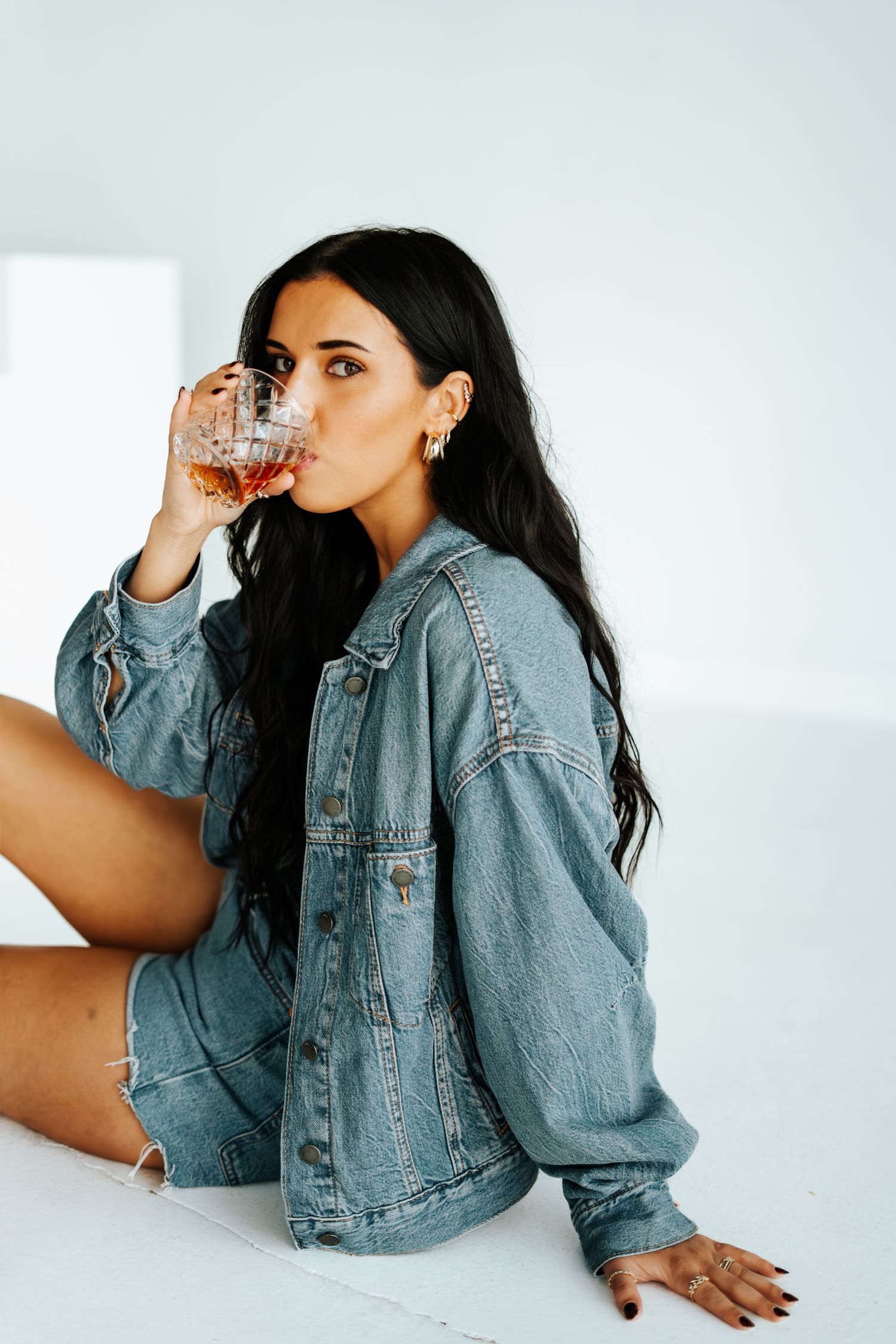 Young brunette woman in oversized denim jacket drinking whiskey while sitting on white floor.
