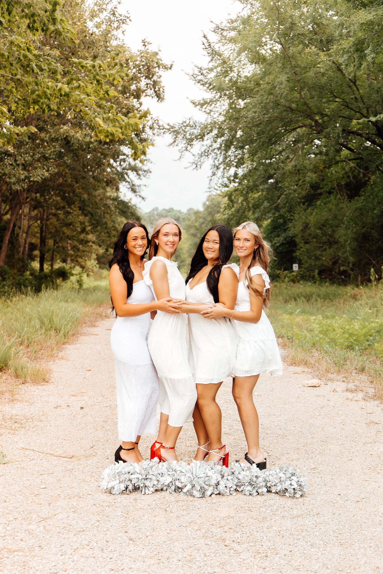 Four young women in white dresses pose together on a dirt path surrounded by trees, holding silver pom-poms.