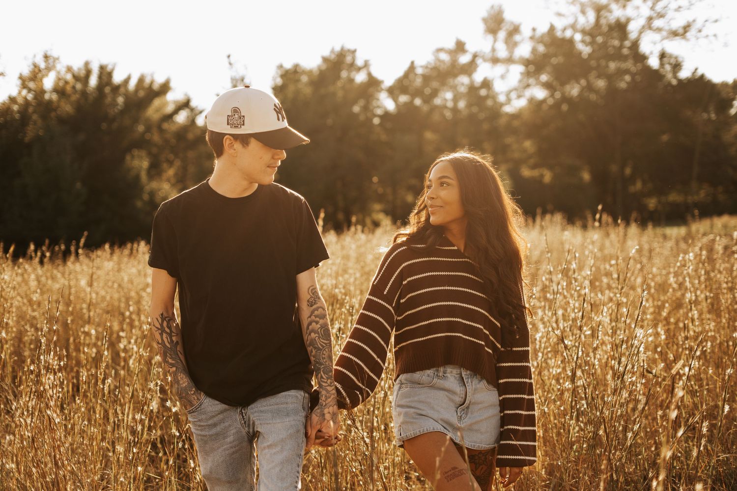 Young couple holds hands walking through golden sunlit field of tall grass at sunset.