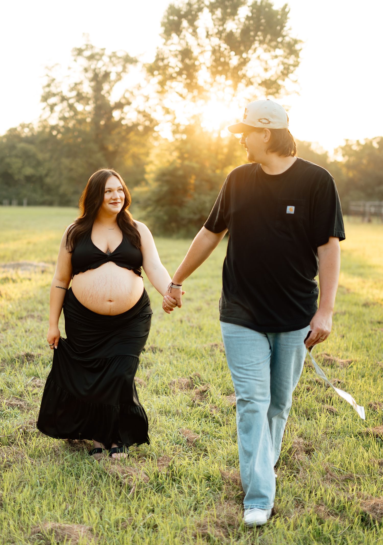 Pregnant woman in black dress holding hands with partner walking in sunlit field.