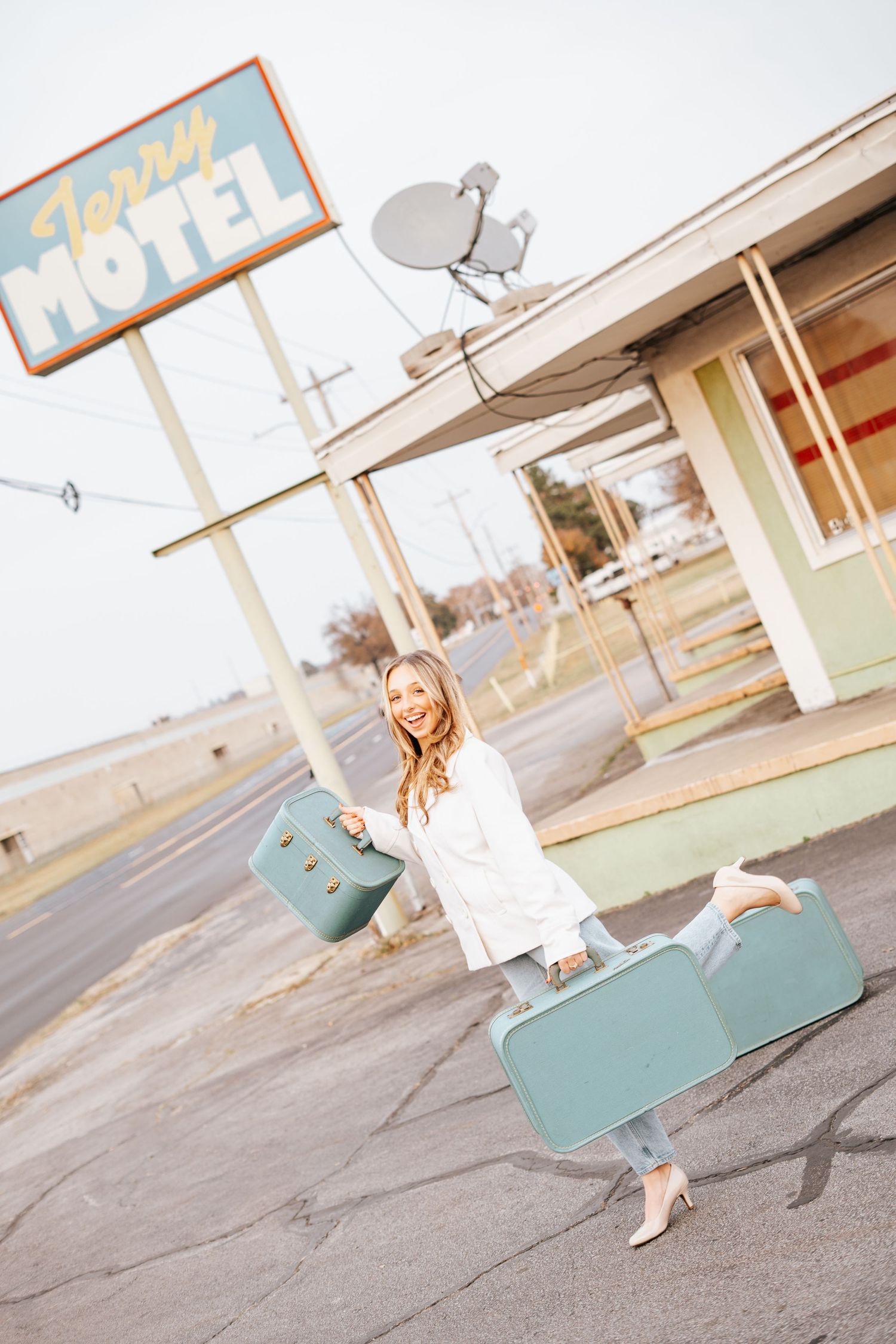 Smiling blonde woman carrying two vintage teal suitcases outside a retro motel on a cloudy day.