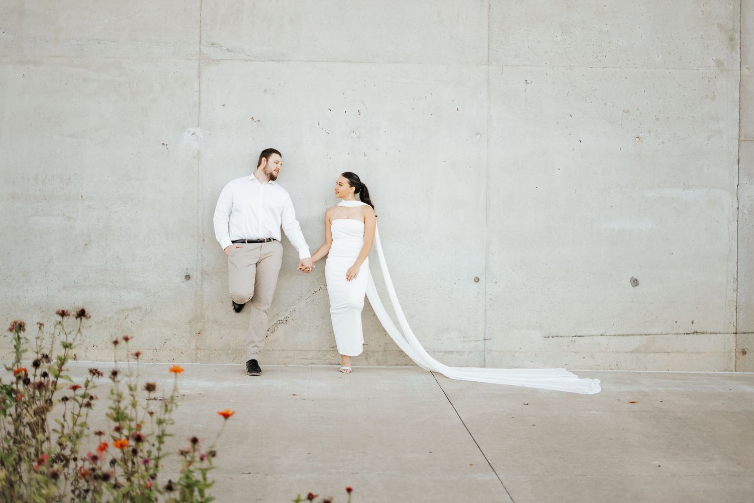 Bride and groom holding hands in front of concrete wall with long flowing veil and colorful wildflowers.