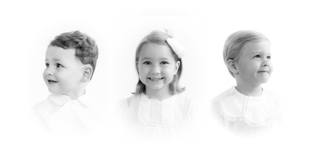 Black and white portrait of three young children in white outfits, smiling and looking sideways.