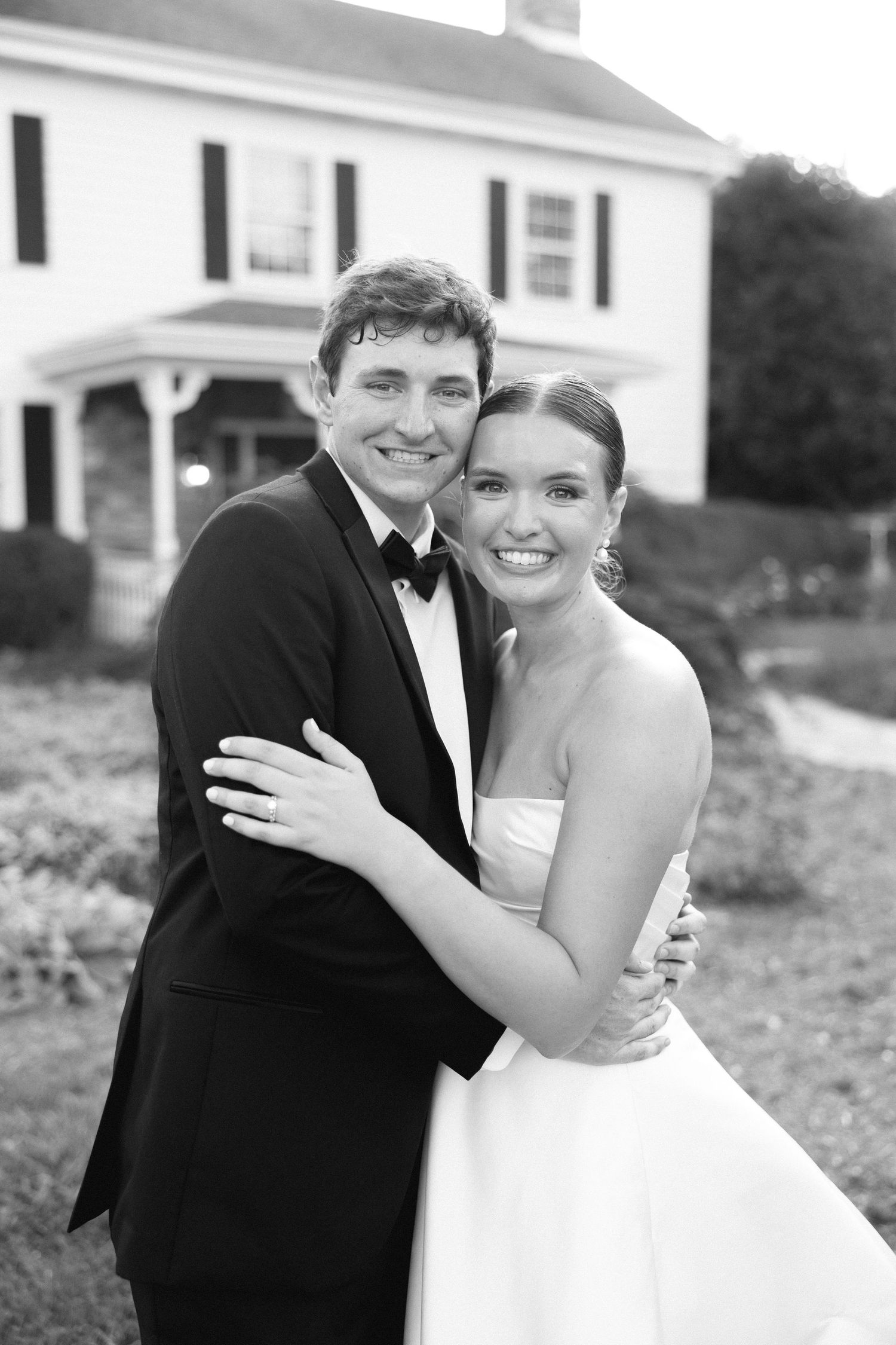 Smiling bride and groom embracing in front of white colonial home in black and white.