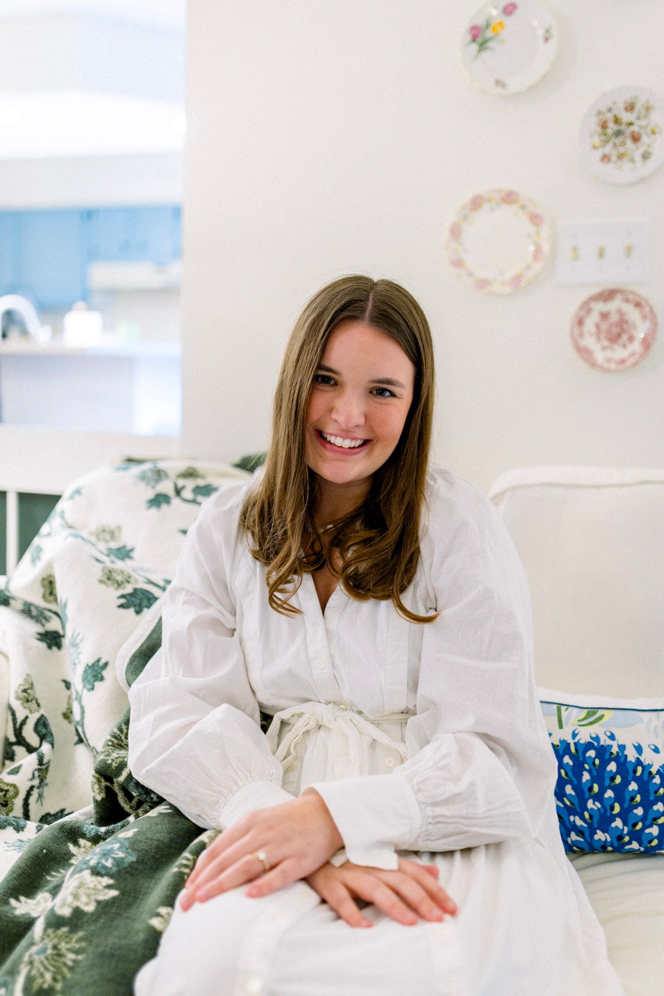 Smiling young woman in a white robe sitting on a bed with floral decor and decorative plates on wall.