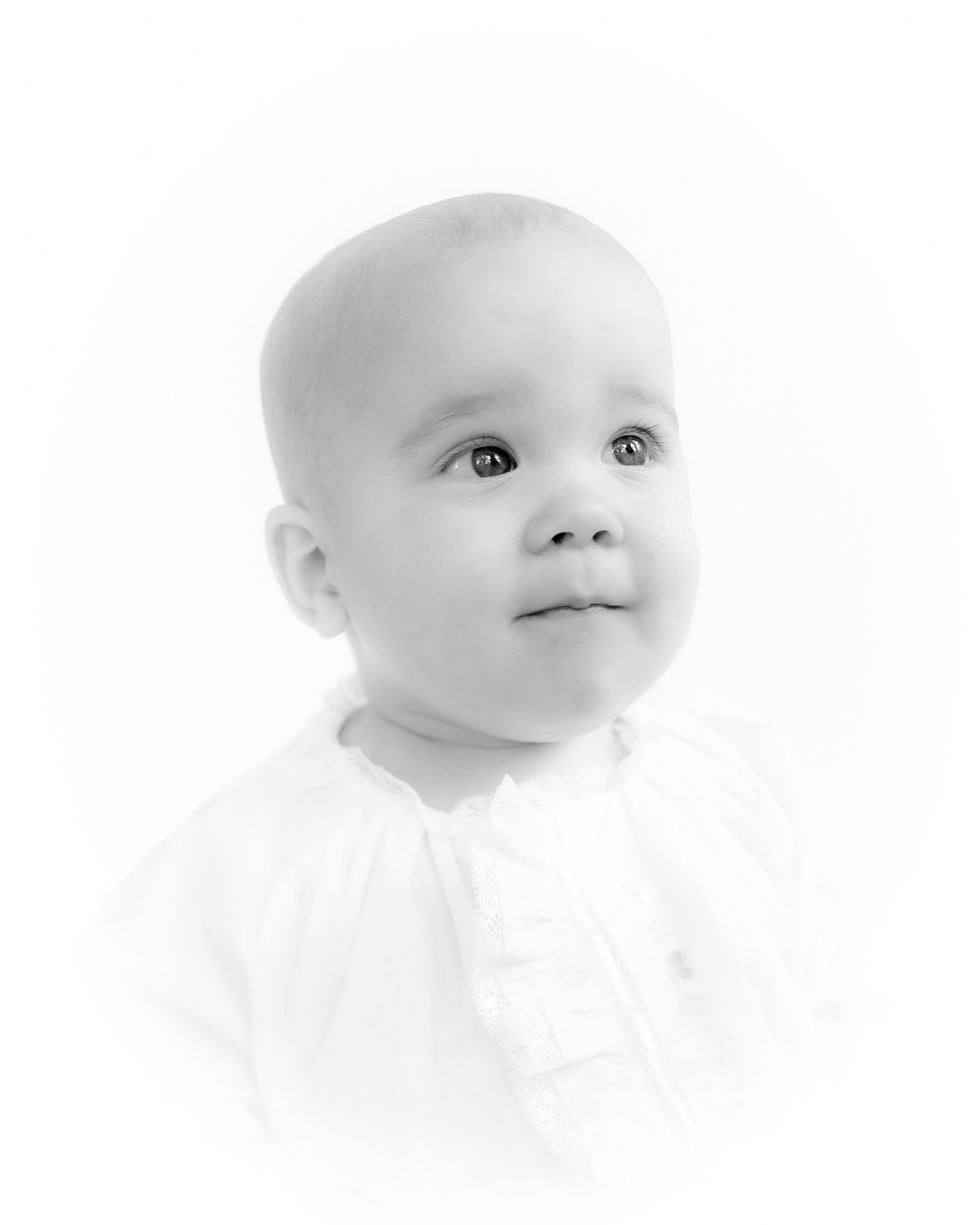 Serene baby in white ruffled outfit looking up, soft black and white studio portrait.