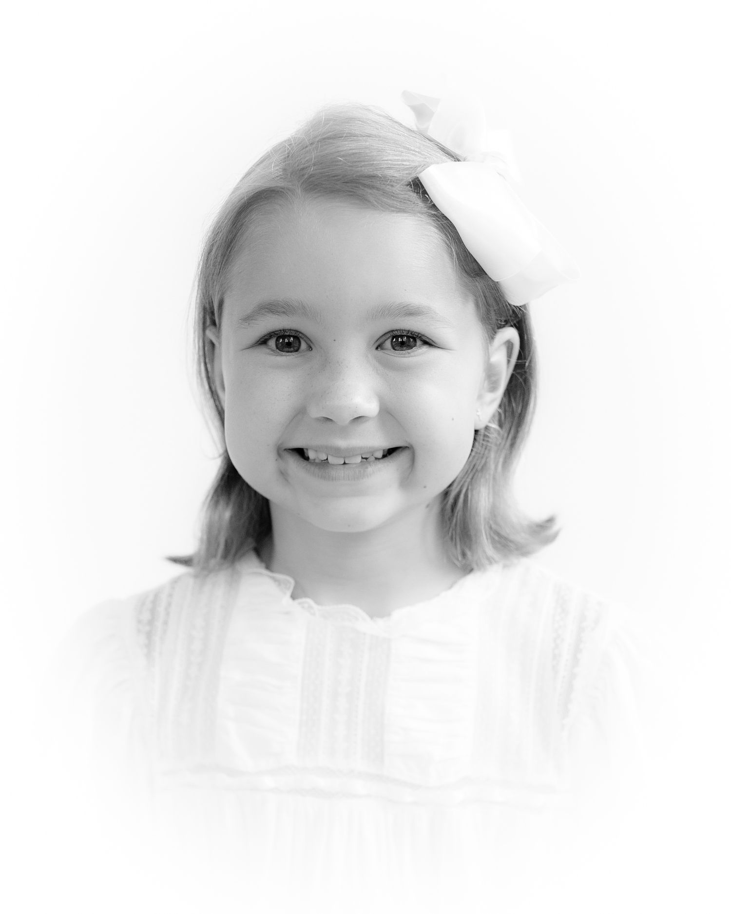 Smiling young girl with white bow, shoulder-length hair, black and white portrait photo.