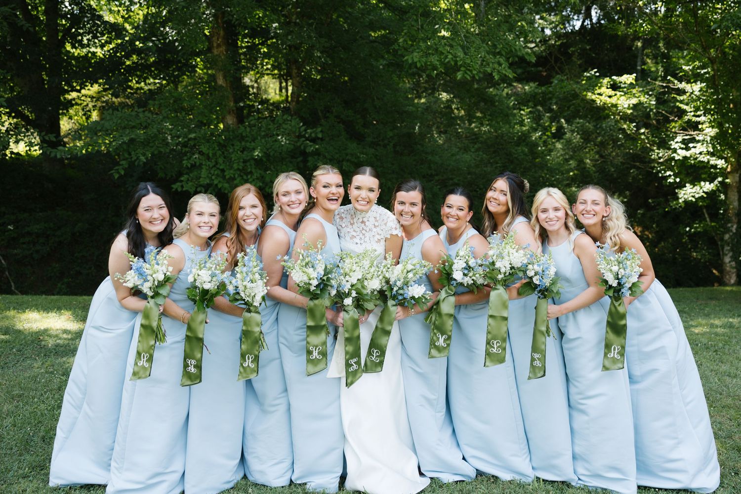 Bride with bridesmaids in light blue gowns holding white floral bouquets outdoors.