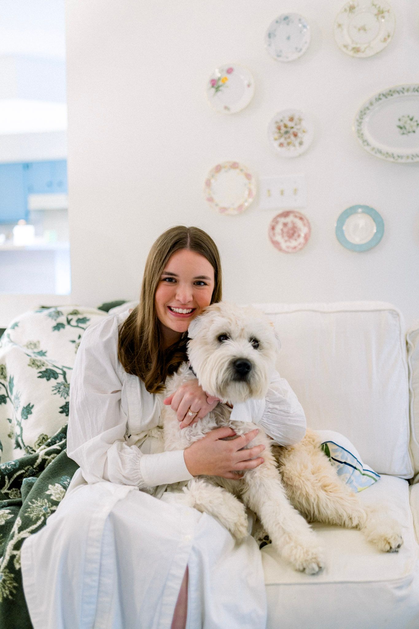 Smiling young woman cuddling white fluffy dog on white sofa in bright home interior.