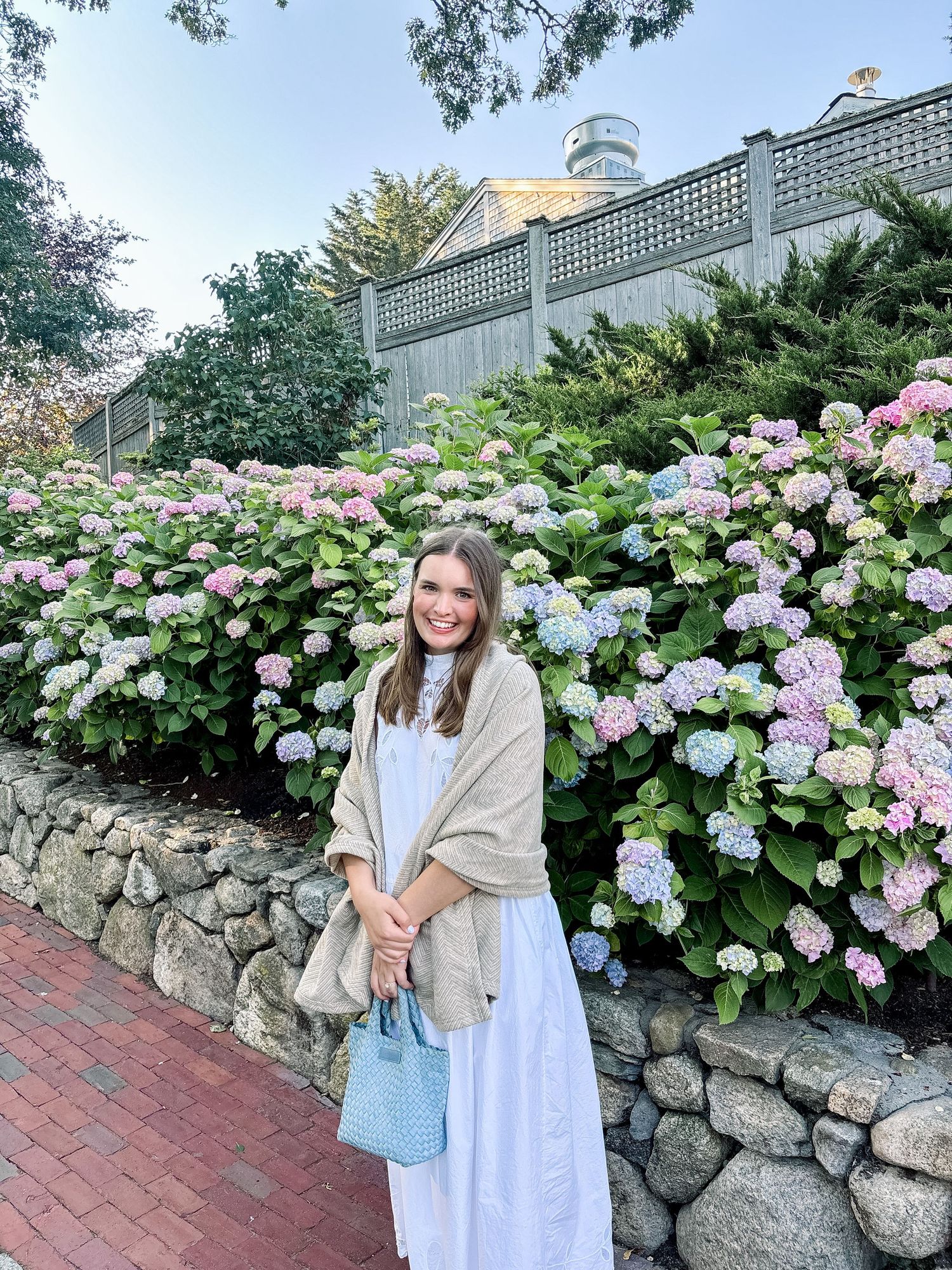 Smiling woman in white dress with shawl standing before blooming hydrangea garden.