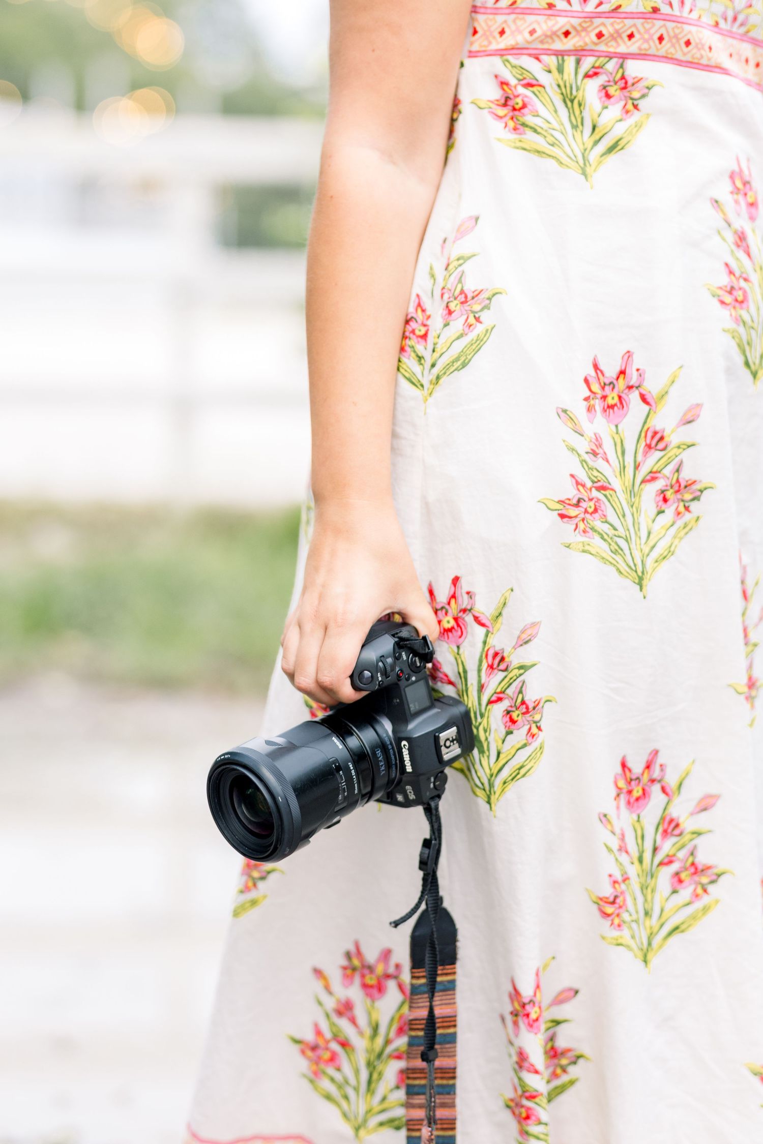 Close-up of a woman in a floral dress holding a DSLR camera with a colorful strap.