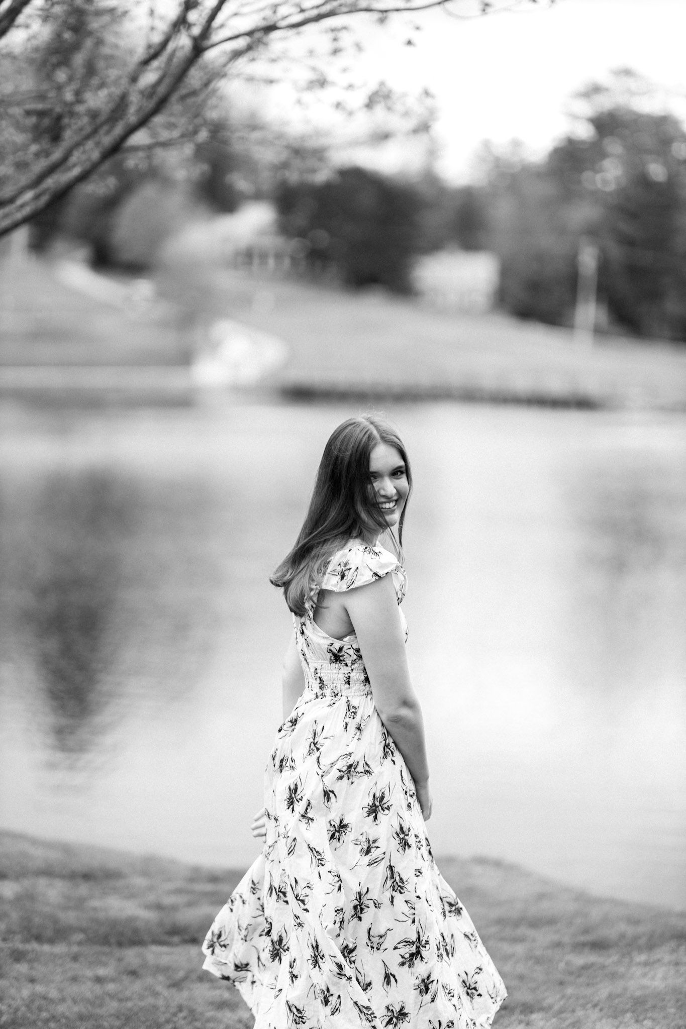 Young woman in floral dress twirling near a peaceful lake in black and white.