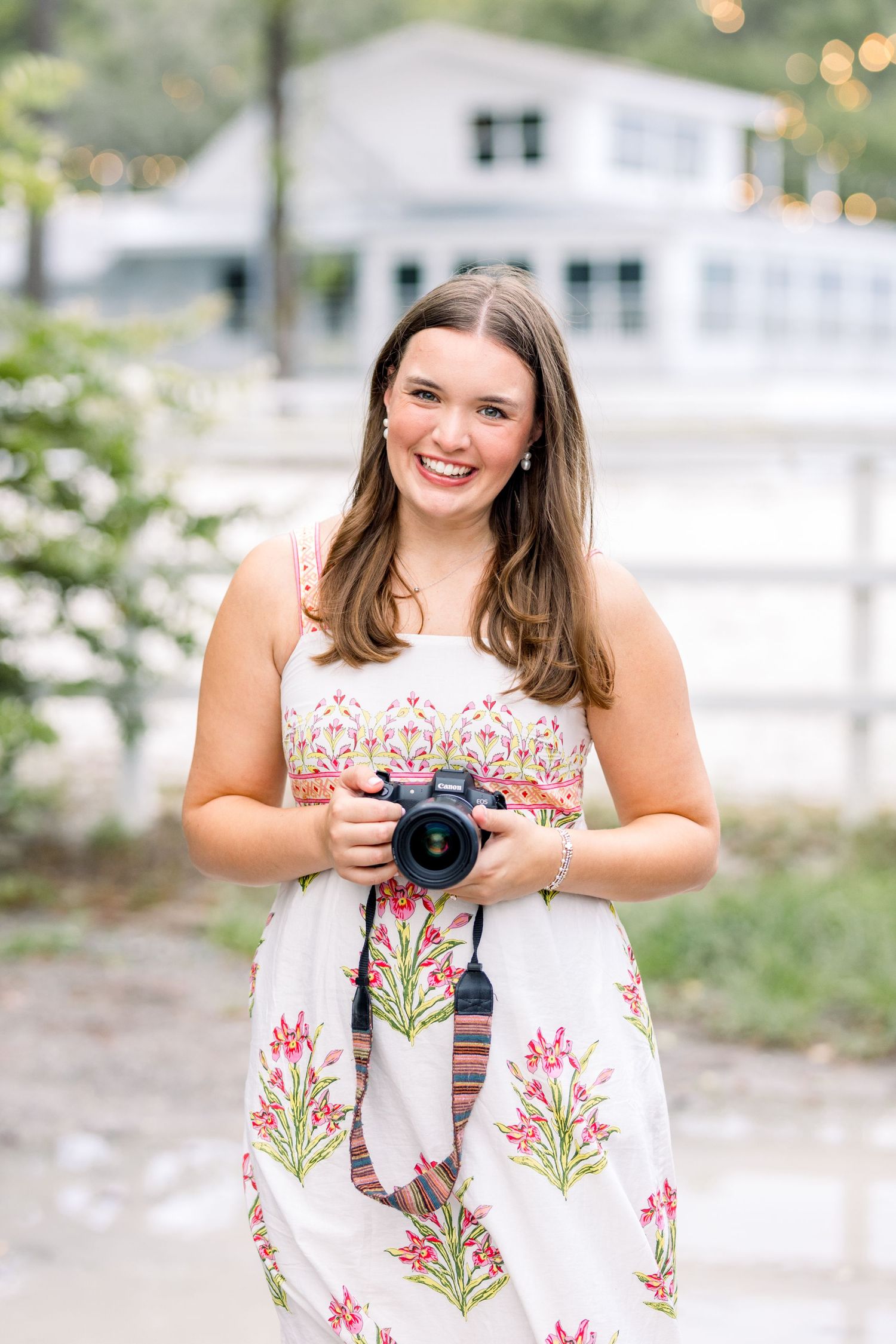 Smiling young woman in a floral dress holding a DSLR camera outdoors near a white building.