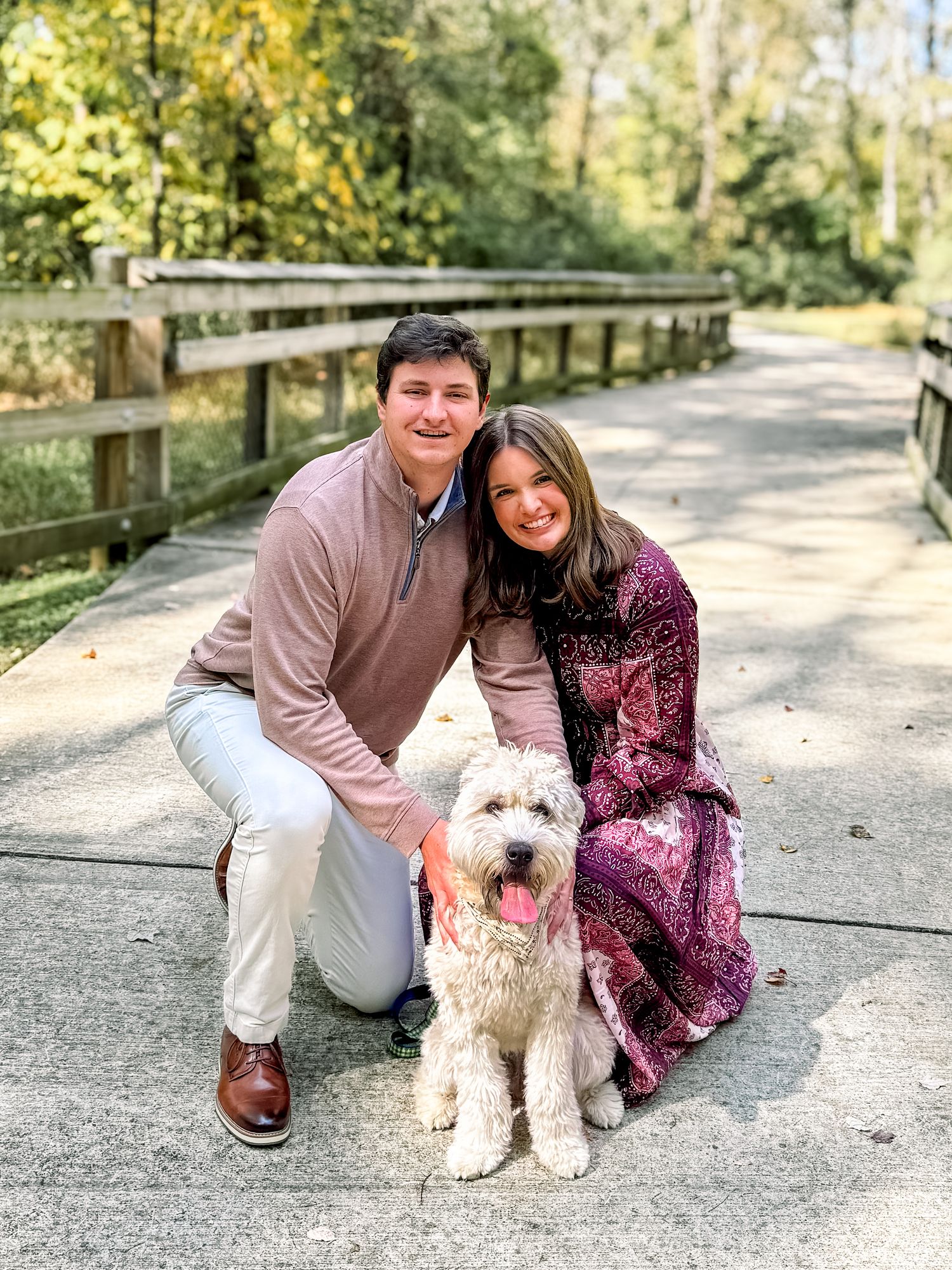 Happy couple posing with fluffy white dog on a scenic outdoor bridge path.
