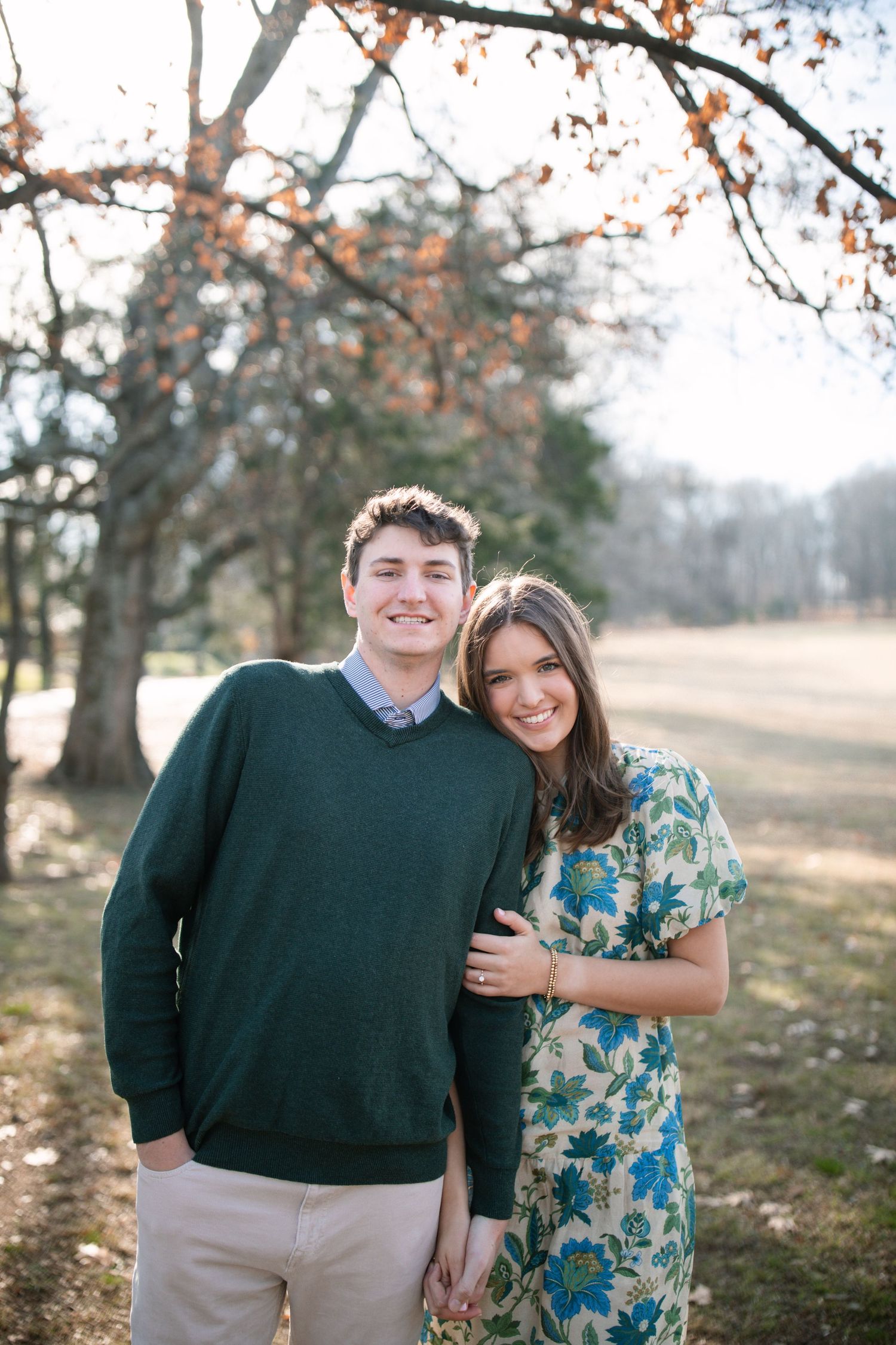 Smiling couple posing outdoors in autumn park, man in green sweater and woman in floral dress.