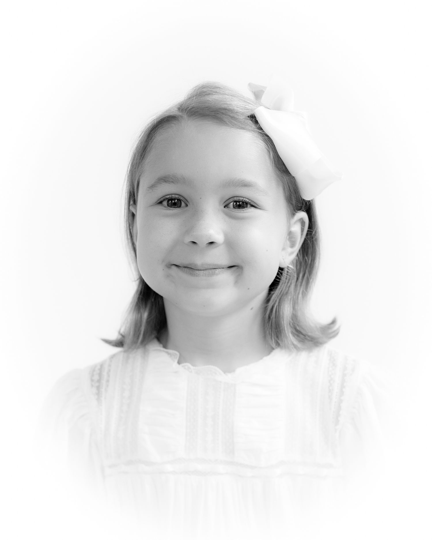 Black and white portrait of a young girl with a bow in her hair, wearing a white dress, smiling softly.