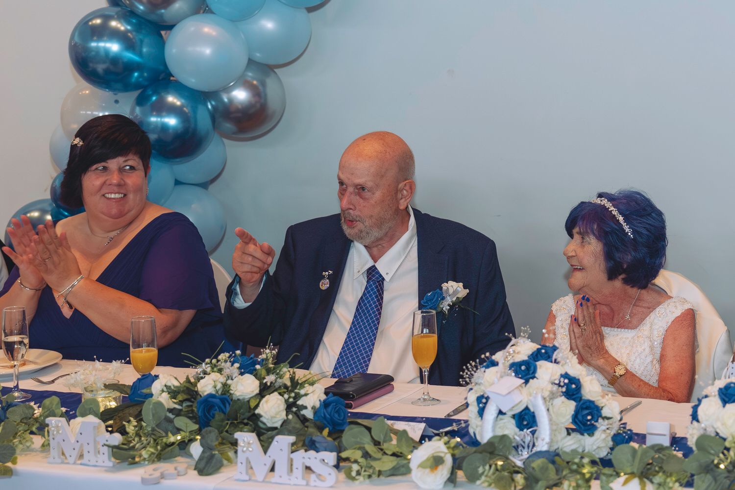 Bride and groom laugh with guest at wedding reception table decorated with blue flowers and Mr & Mrs signs.