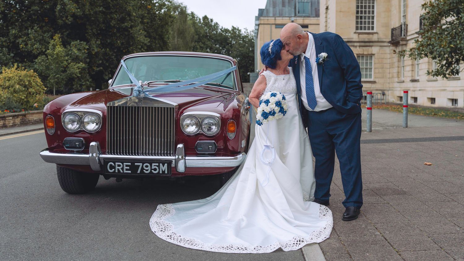 Elderly bride and groom kissing beside a classic burgundy Rolls-Royce wedding car decorated with white ribbons.