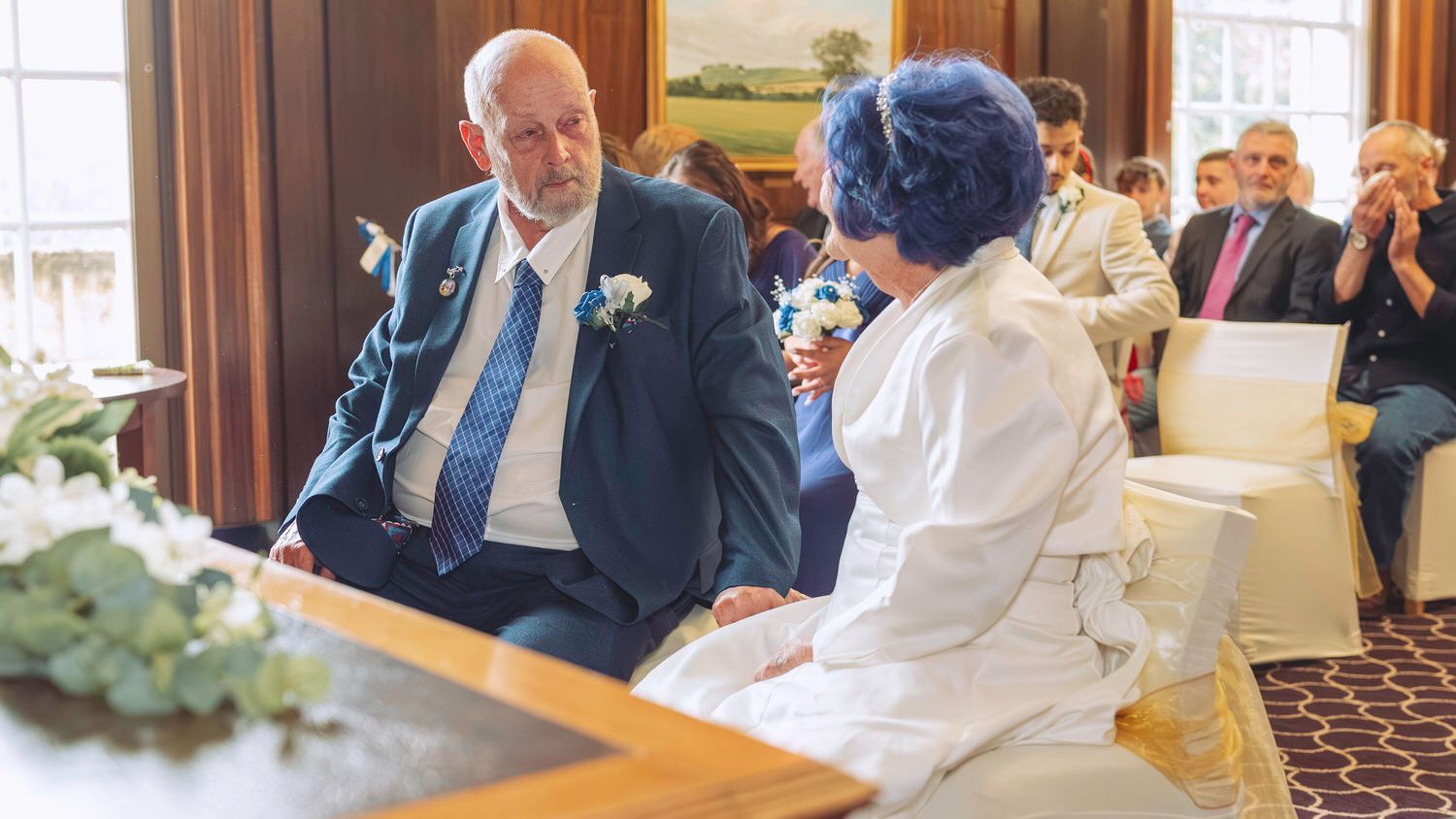 Elderly couple exchanging vows at an indoor wedding ceremony, guests applauding in background.