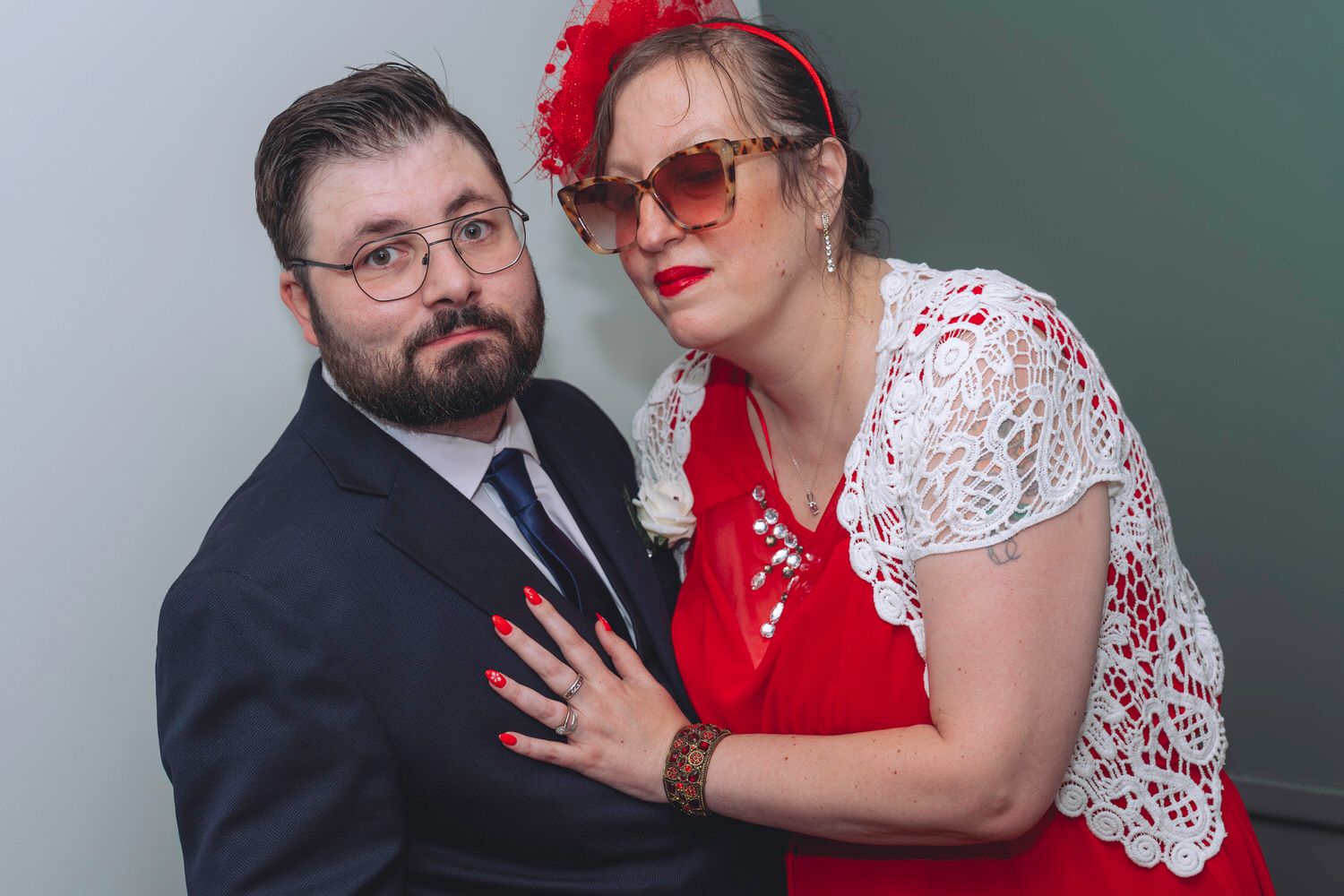 Couple posing together, man in dark suit and woman in red dress with lace overlay and red floral headband.