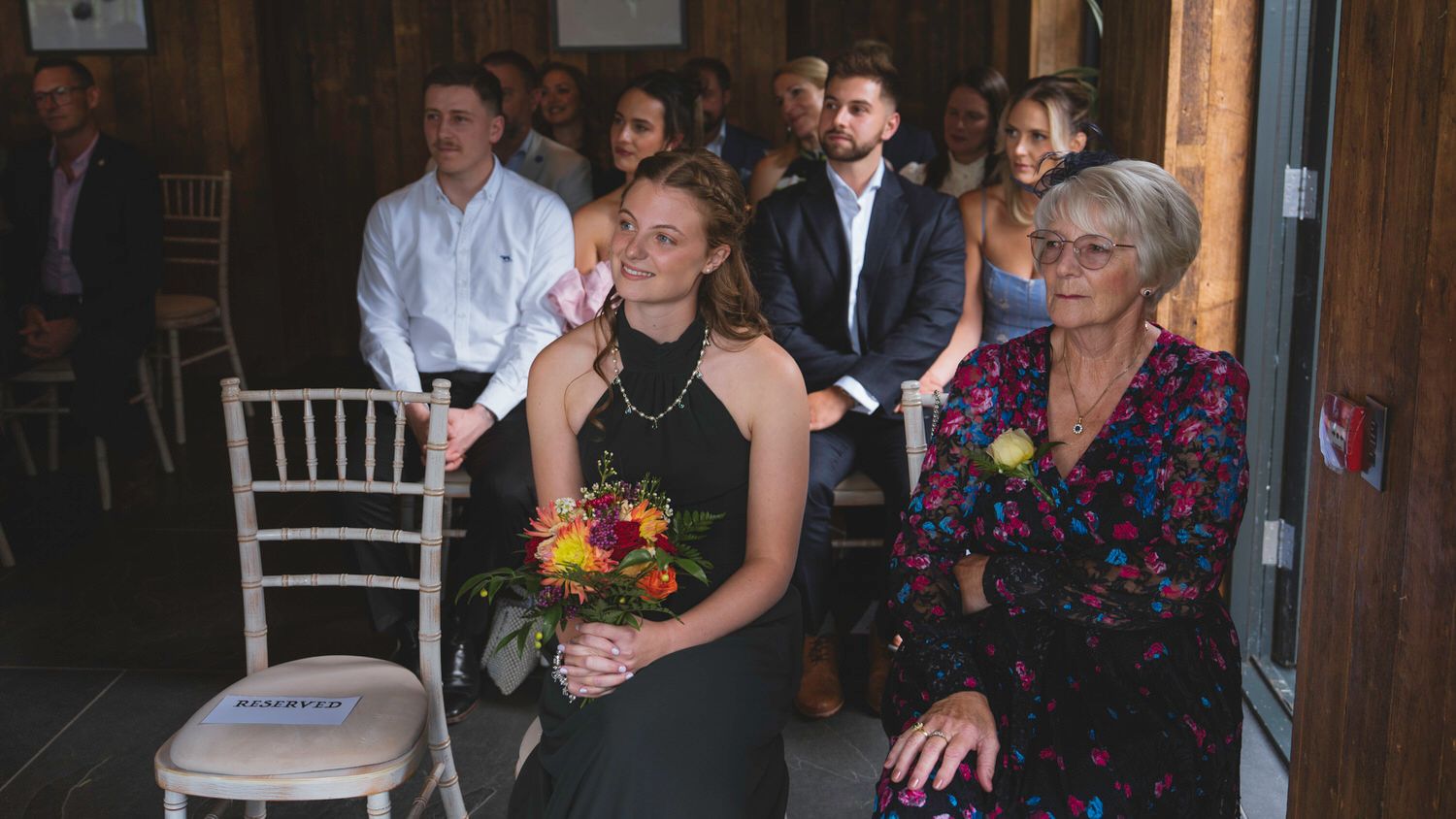 Guests seated at an indoor wedding ceremony, woman in black dress holding colorful bouquet smiling beside older woman in floral top.