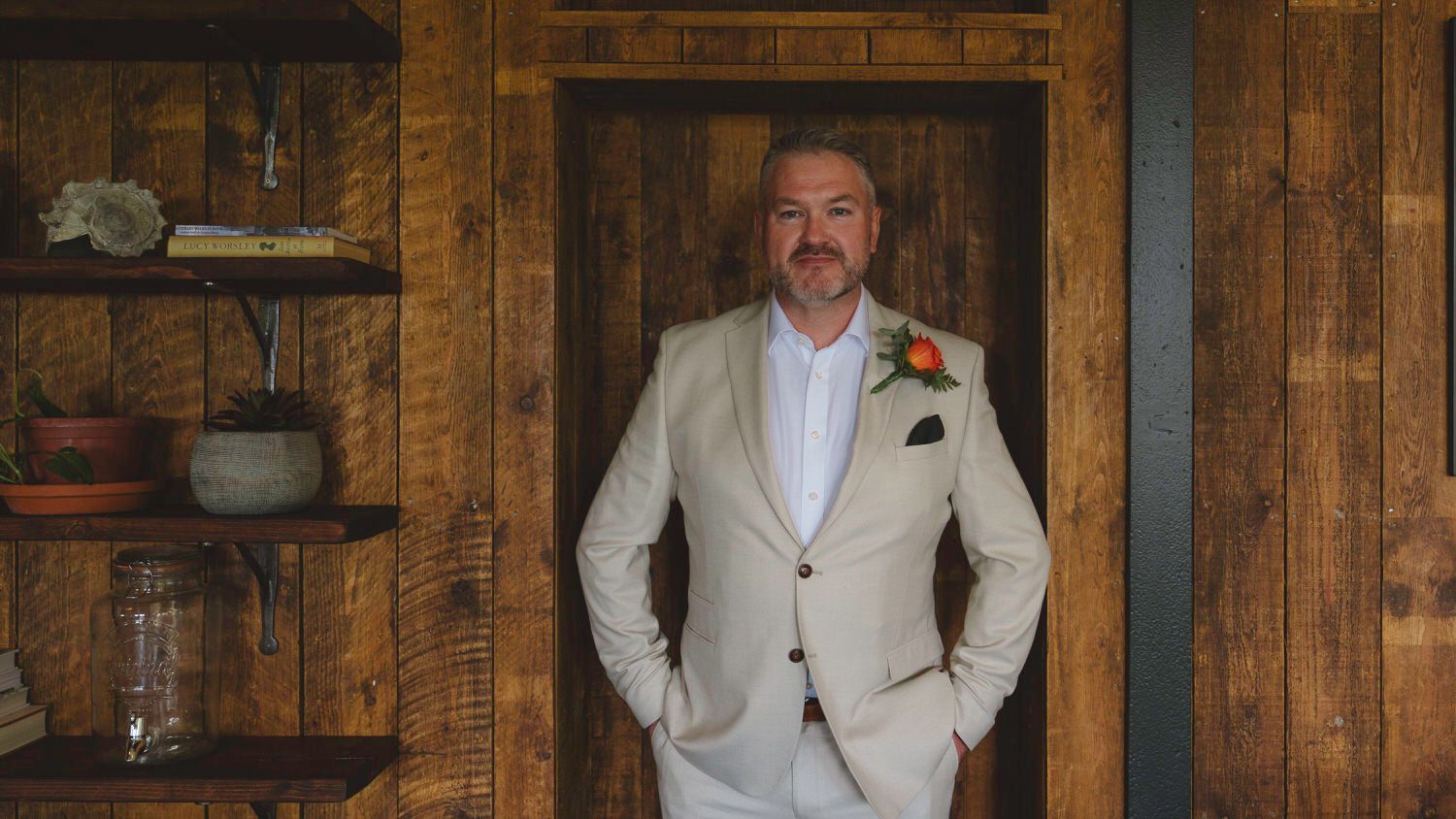 Man in beige suit with boutonniere poses in rustic wooden interior near shelves.