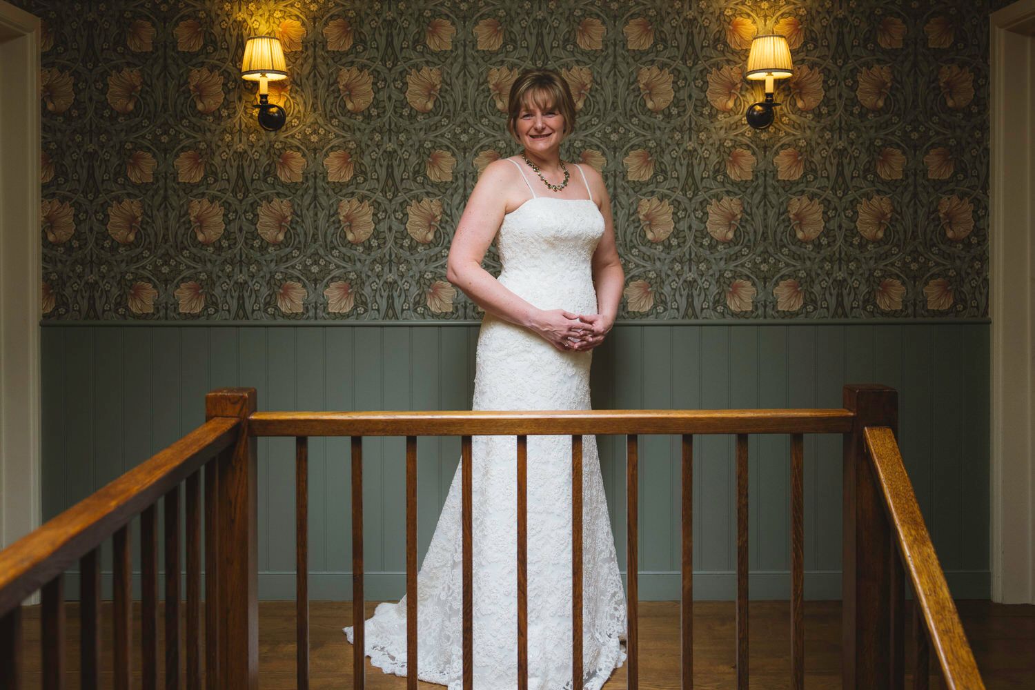 Bride in white lace wedding gown stands at staircase railing with vintage floral wallpaper and wall sconces behind her.