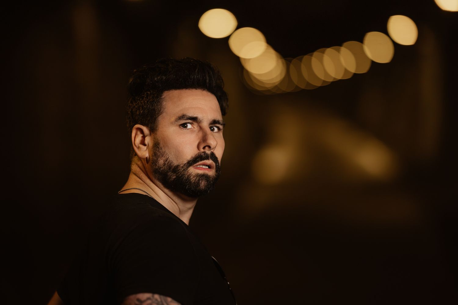Bearded man with tattoos in black t-shirt looking back with bokeh lights background.